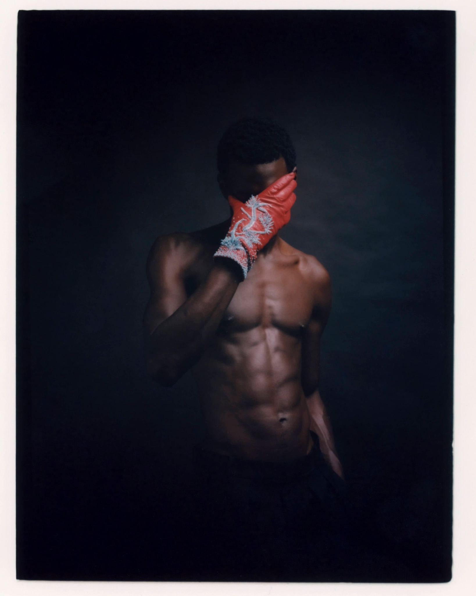 Bare-chested model holding a red-gloved hand over their face against a dark background.