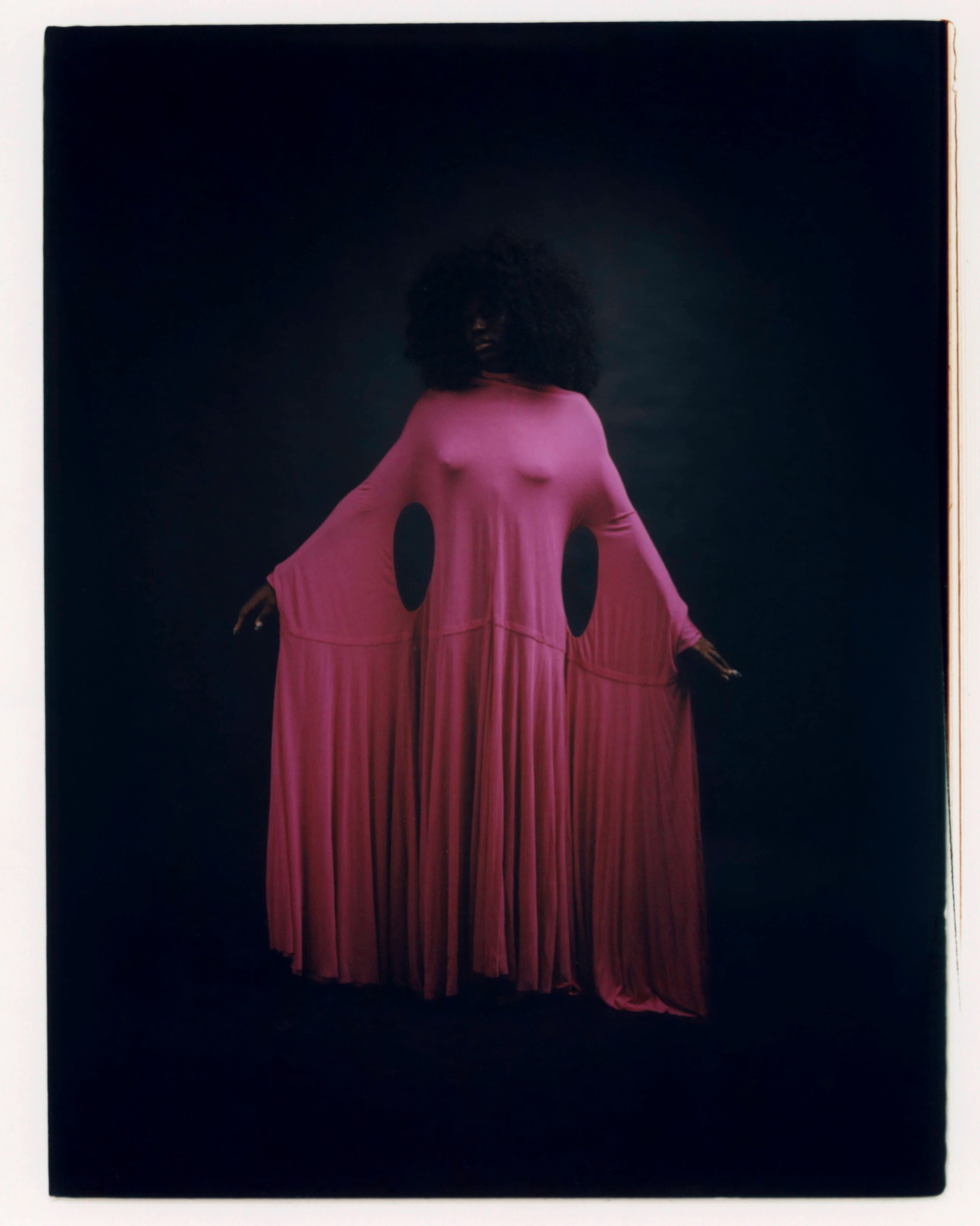 Model seen from behind wearing a vivid pink dress with long hanging strips against a dark studio background.