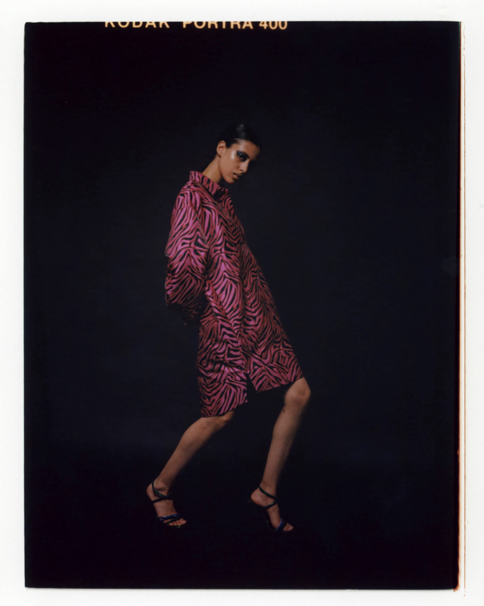 Full-length portrait of a model in a pink patterned dress and heels, posed sideways against a dark backdrop.