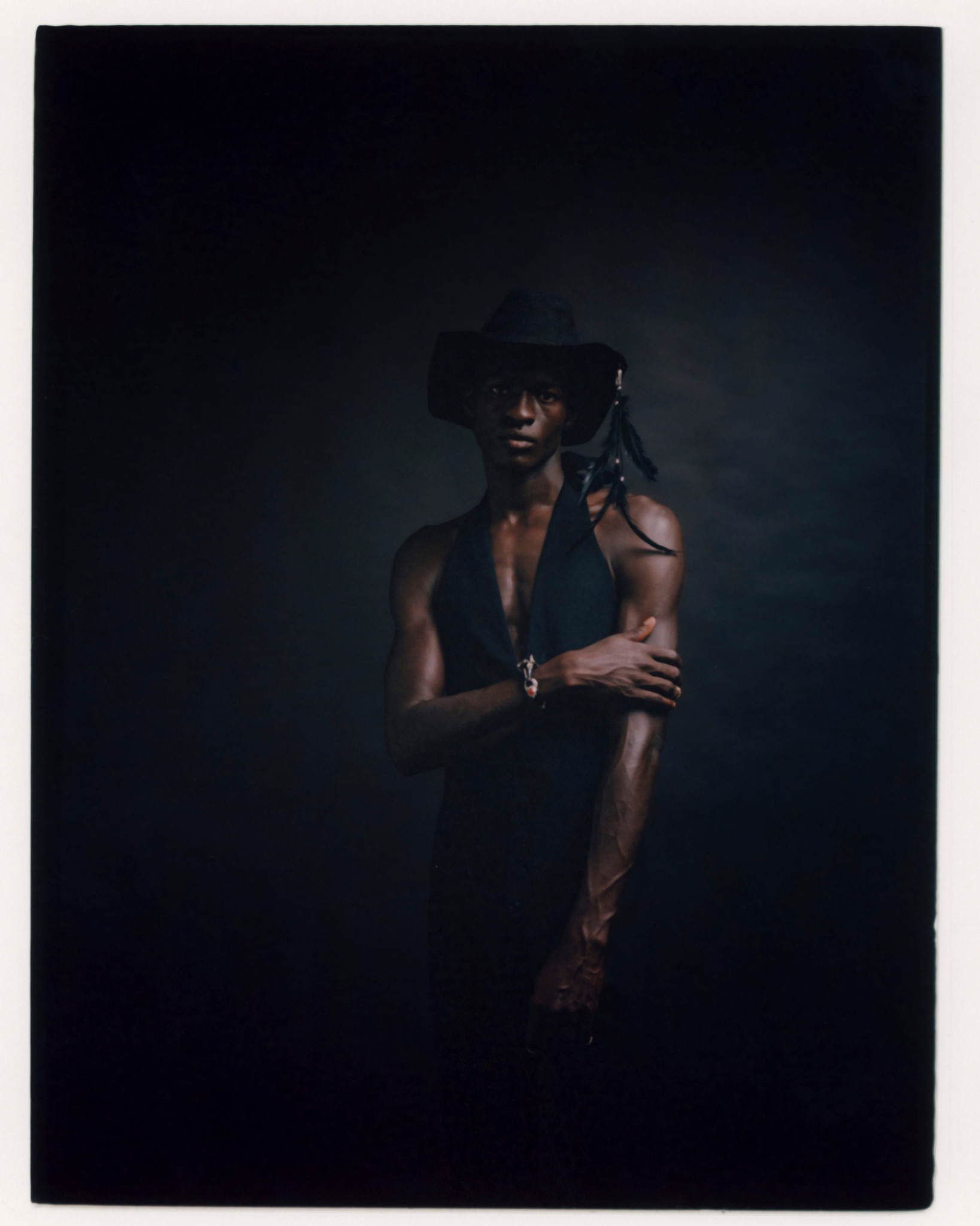 Model in a dark wide-brimmed hat and sleeveless top, holding one arm against a dark backdrop.