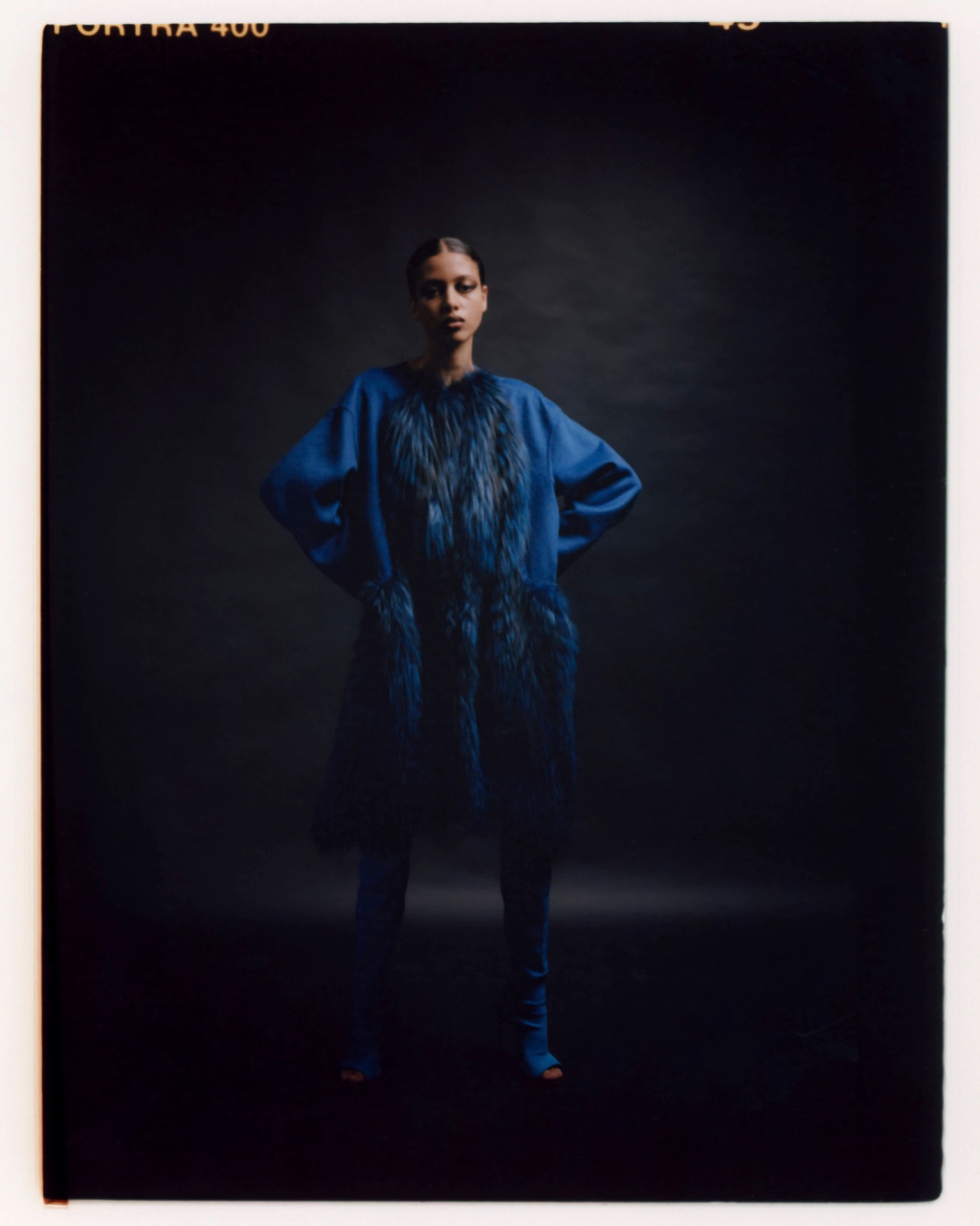 Full-length portrait of a model in a blue outfit with voluminous sleeves, hands on hips against a dark backdrop.