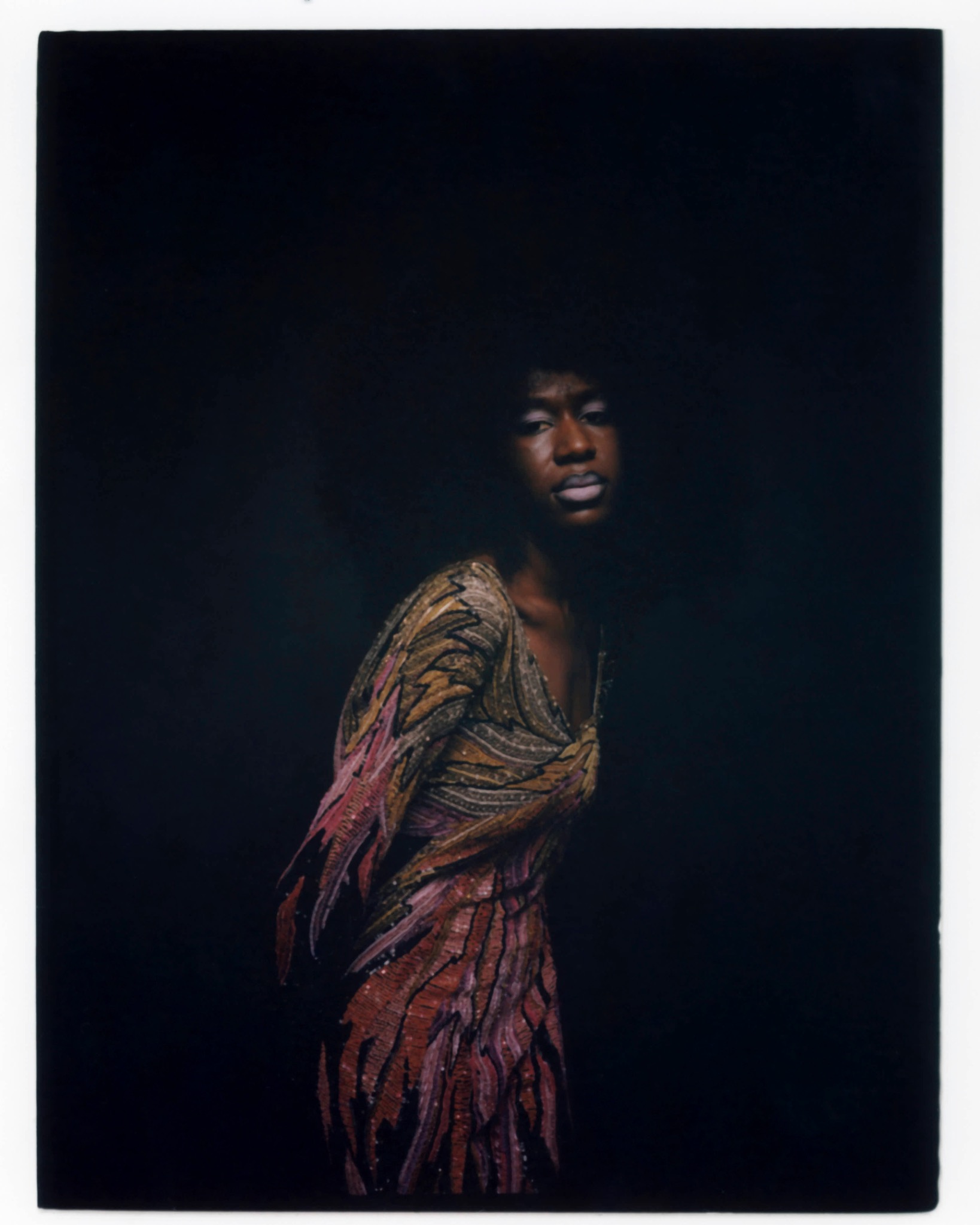 Model turned sideways in a flowing multicoloured patterned outfit against a dark background.