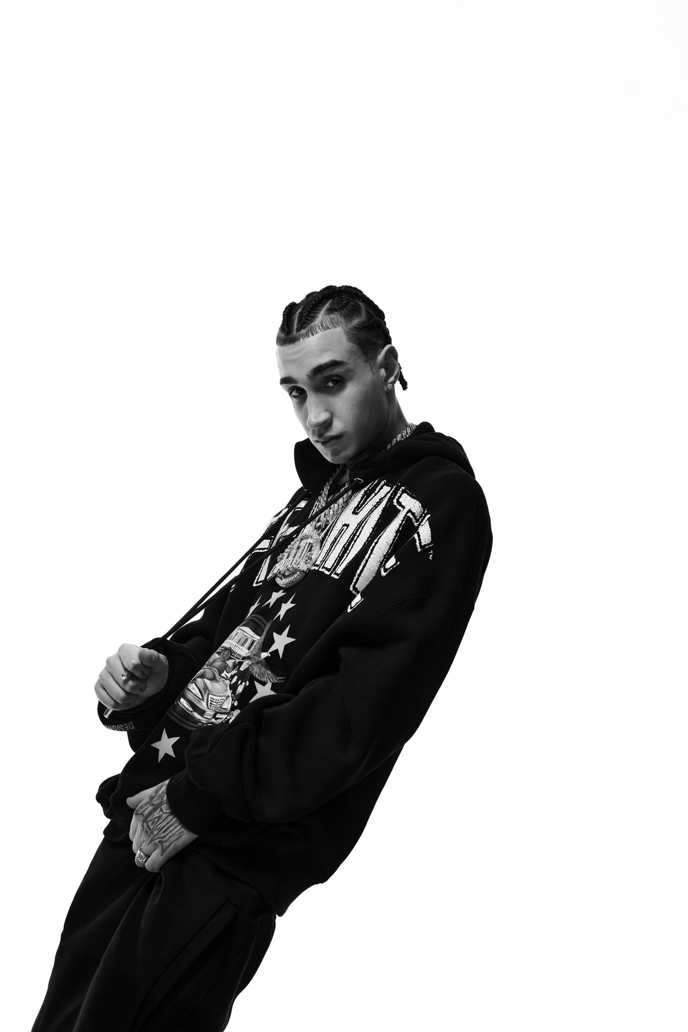 Black-and-white studio portrait of Capo Plaza leaning sideways in a dark graphic sweatshirt.