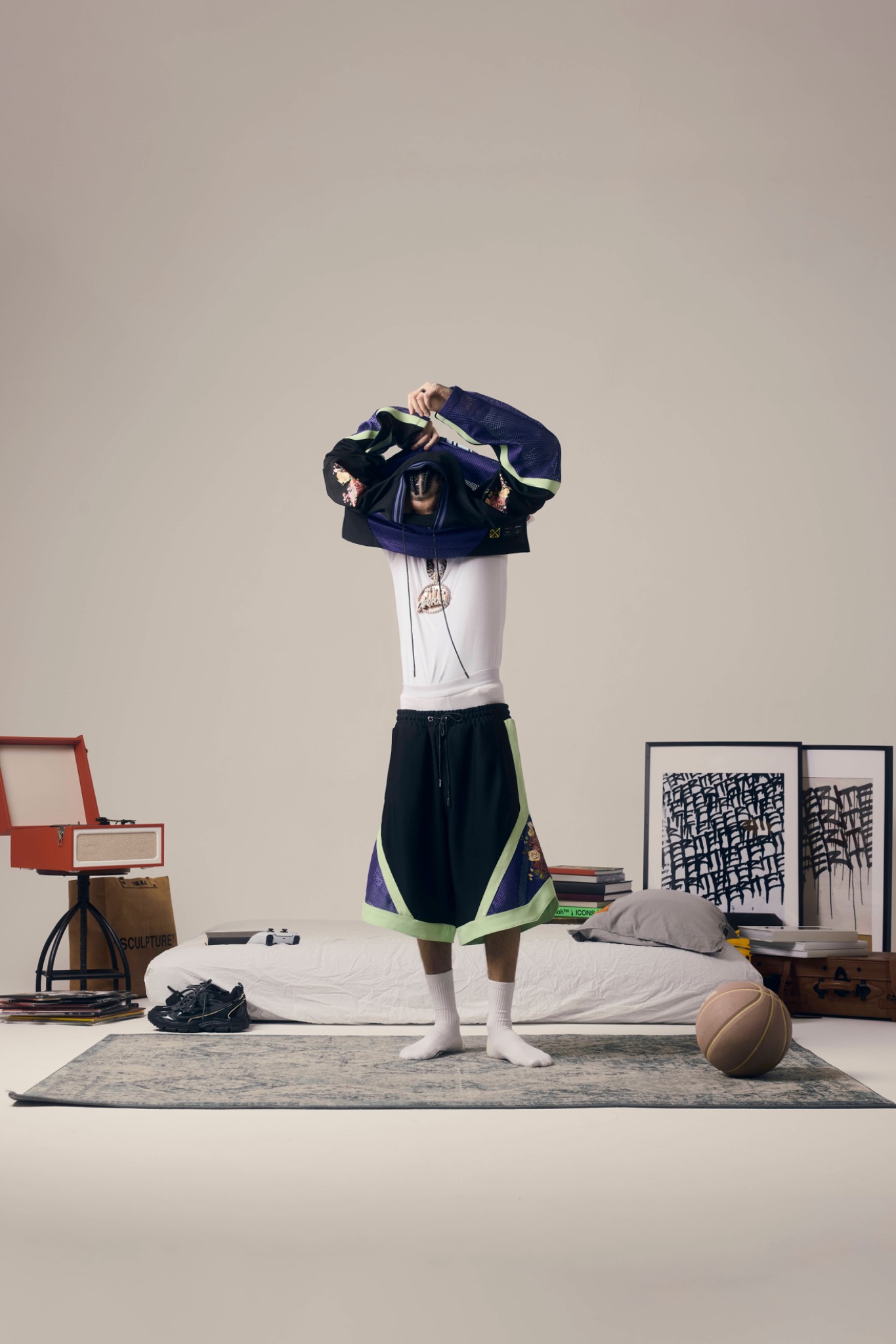 Model pulling a dark patterned top over their head, standing among framed pictures and furniture in a studio.