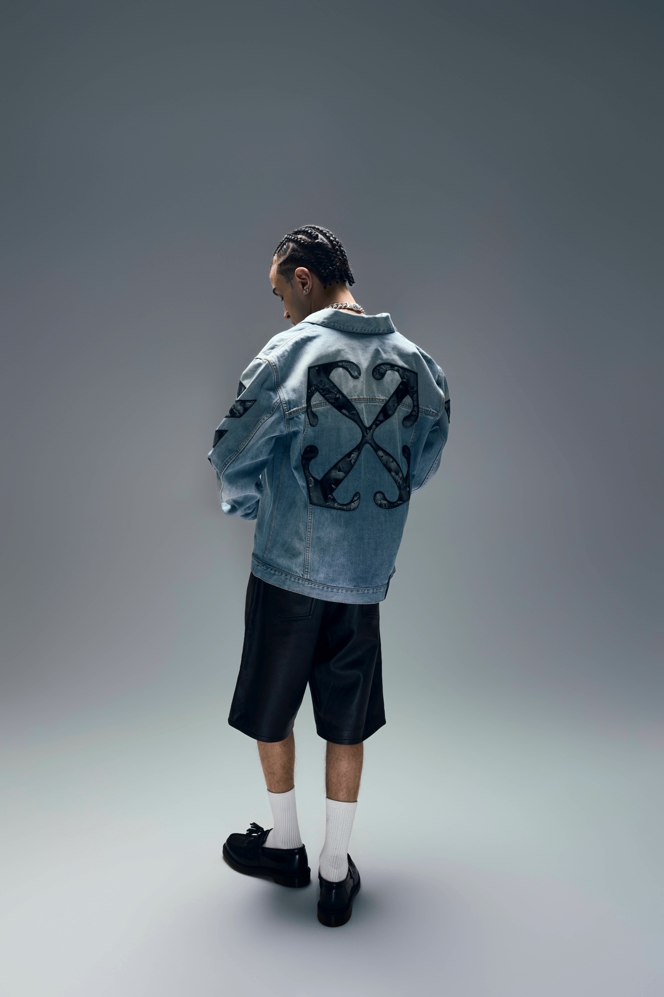 Capo Plaza seen from behind in a denim jacket with a large crossed-arrow motif, dark shorts and white socks.