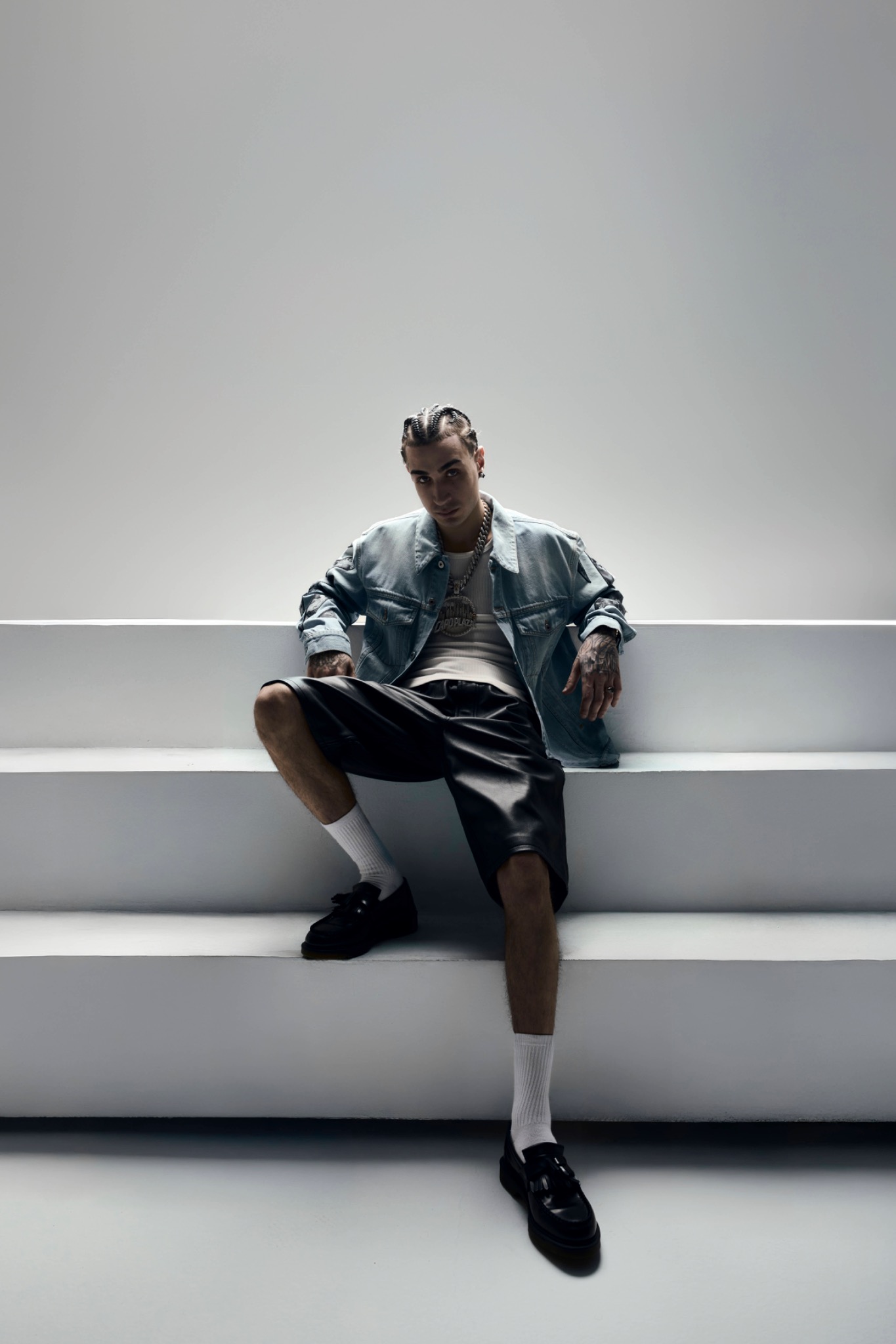 Full-length studio portrait of Capo Plaza seated on white steps in a denim jacket, shorts and dark shoes.