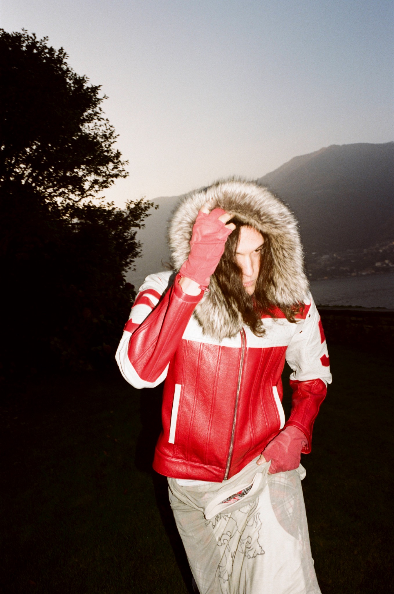 Model in a red jacket with a fur-trimmed hood, holding a red-gloved hand against their face by a lake.