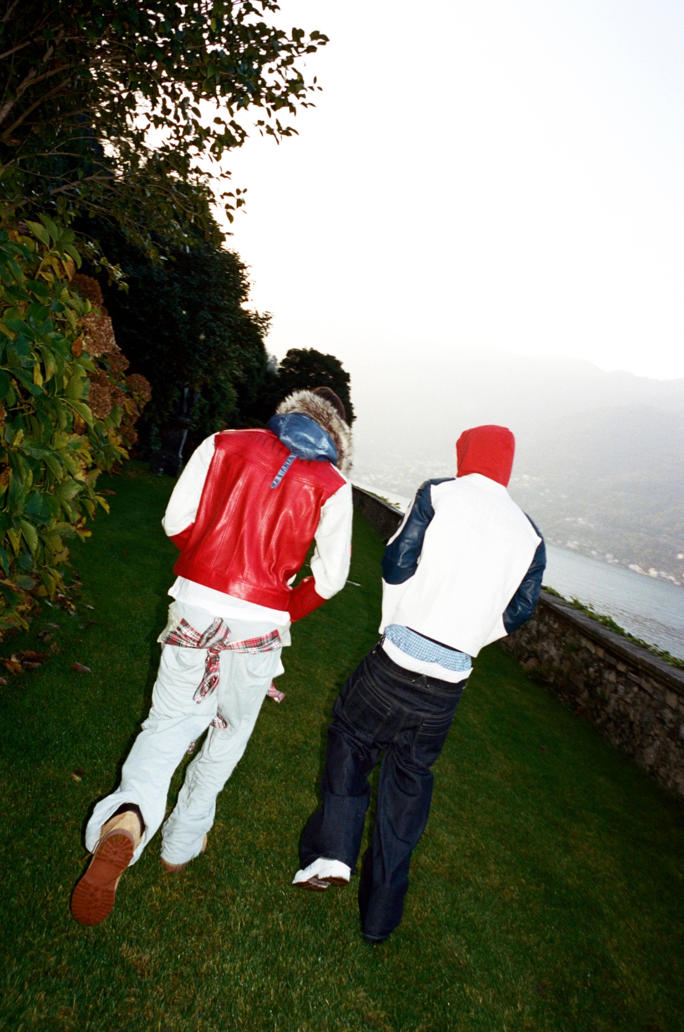 Two models in red, white and blue jackets running across grass beside a lake.
