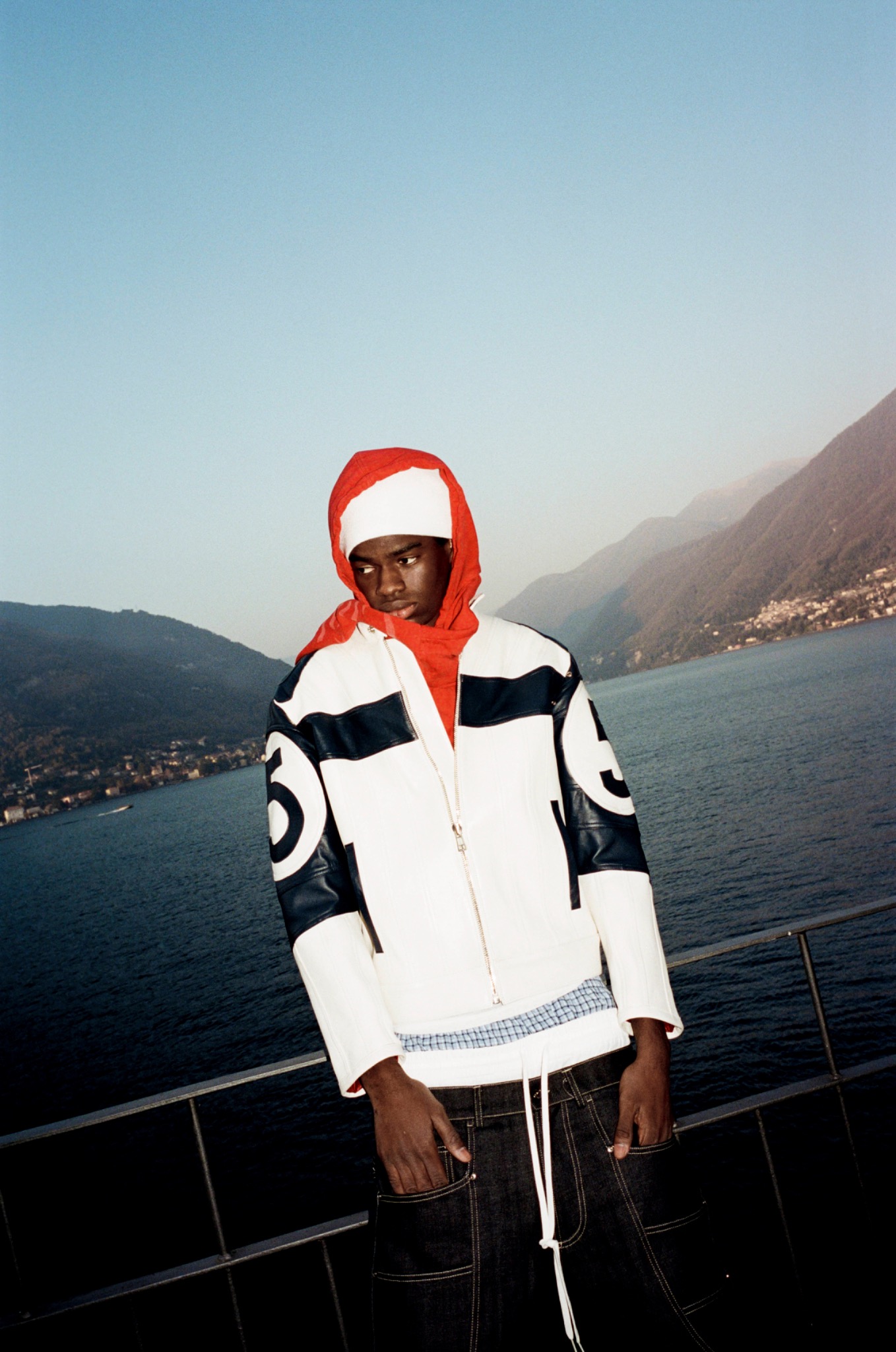 Model in a white patterned jacket with a red hood standing by a lakeside railing, mountains behind.