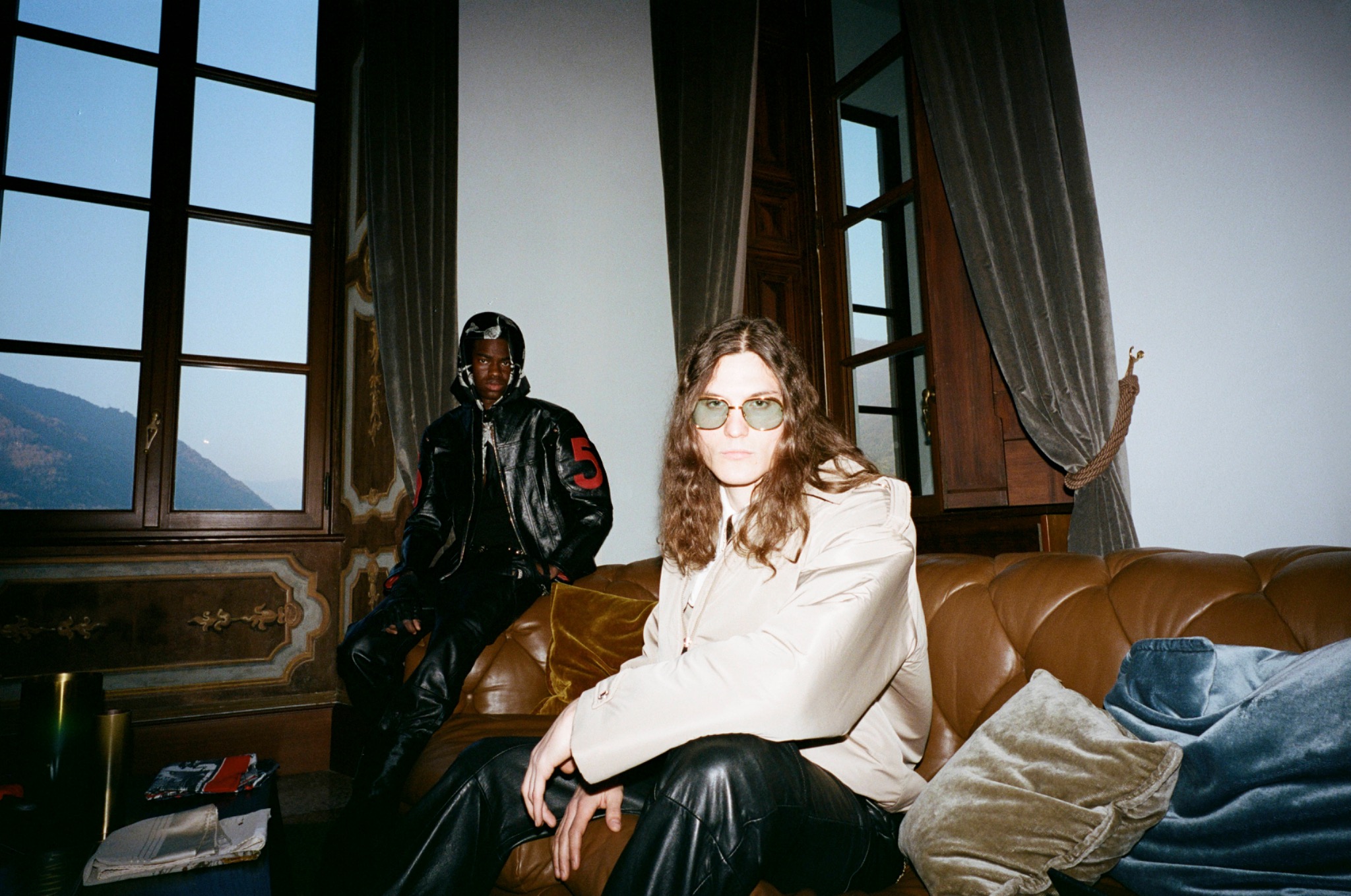 Two models in sunglasses inside a room with tall windows, one seated in the foreground in a pale top.
