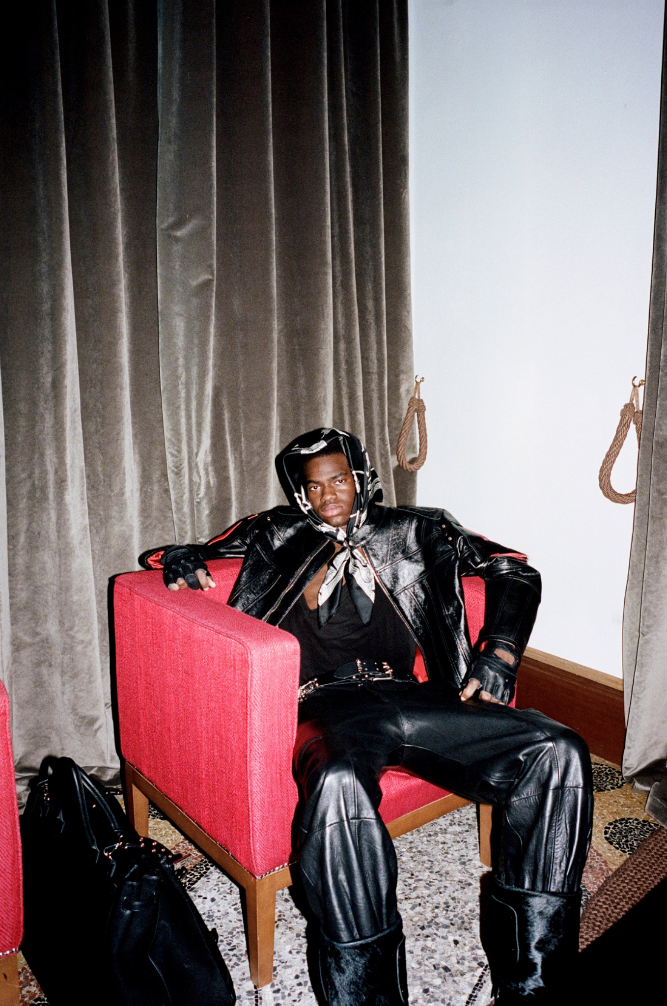 Model in a black leather outfit and cap sitting in a red armchair beside dark curtains.