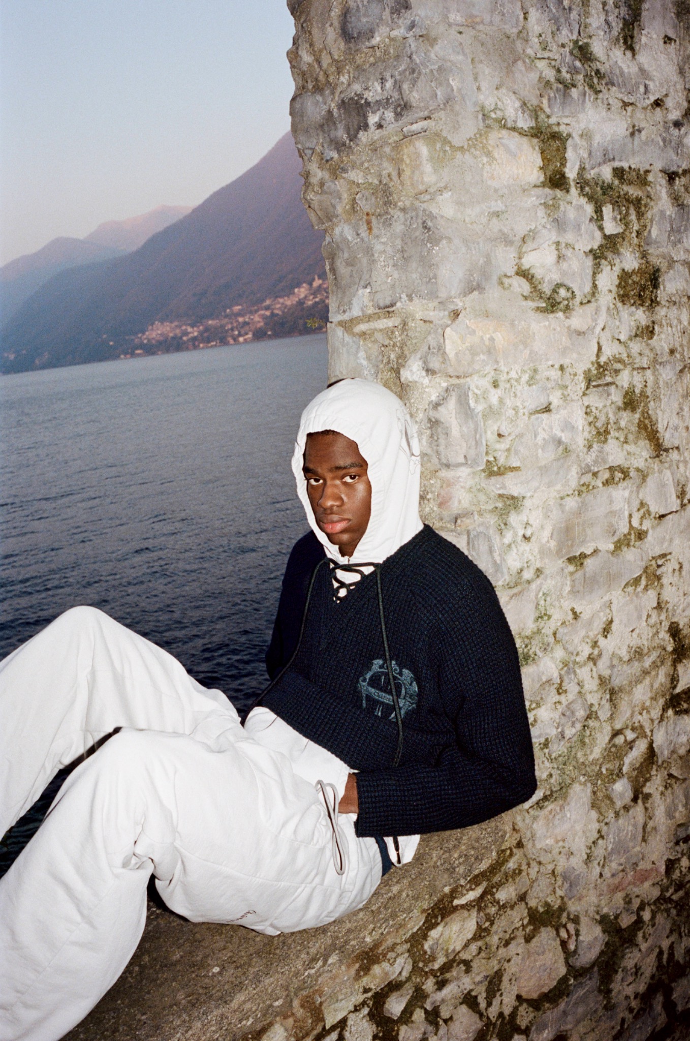 Model in a dark hoodie and white trousers seated against a stone wall beside a lake.