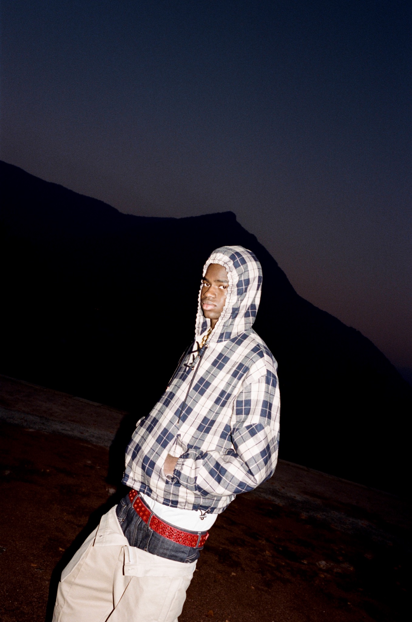 Model in a checked hooded jacket photographed outdoors at dusk, mountains silhouetted behind.