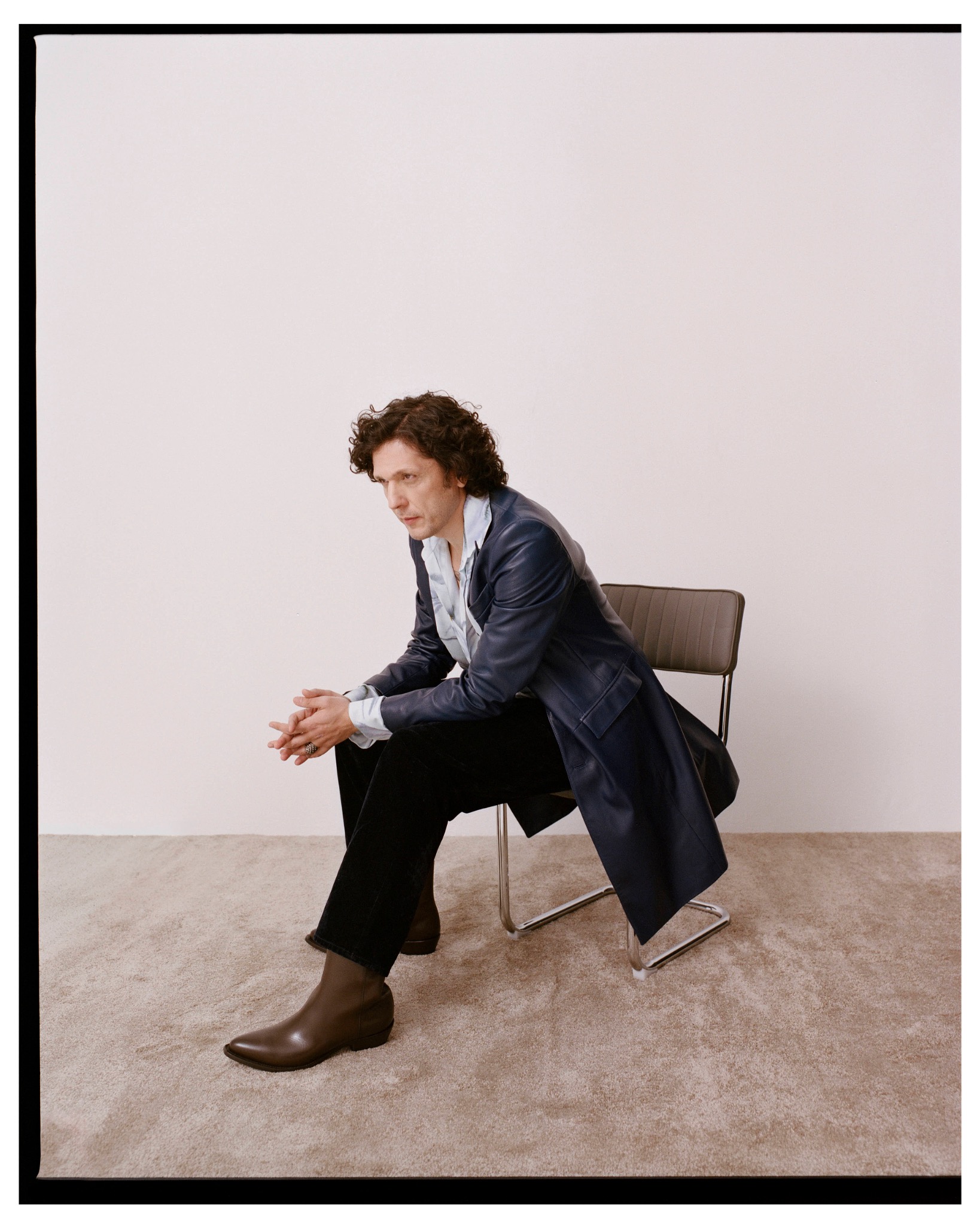 Ermal Meta leaning forward on a chair in a dark coat, cropped trousers and brown boots.