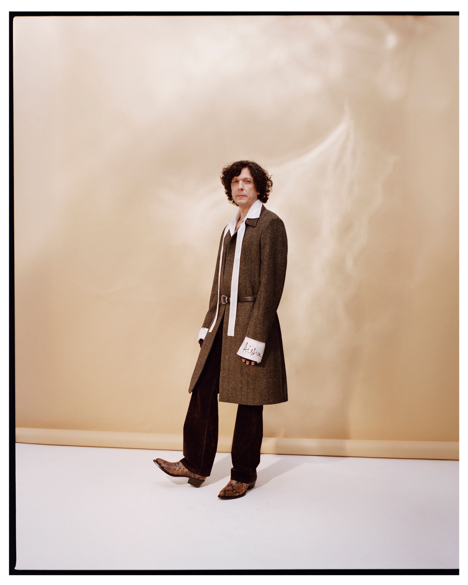 Full-length portrait of Ermal Meta in a long brown coat, white shirt and tie against a pale studio backdrop.