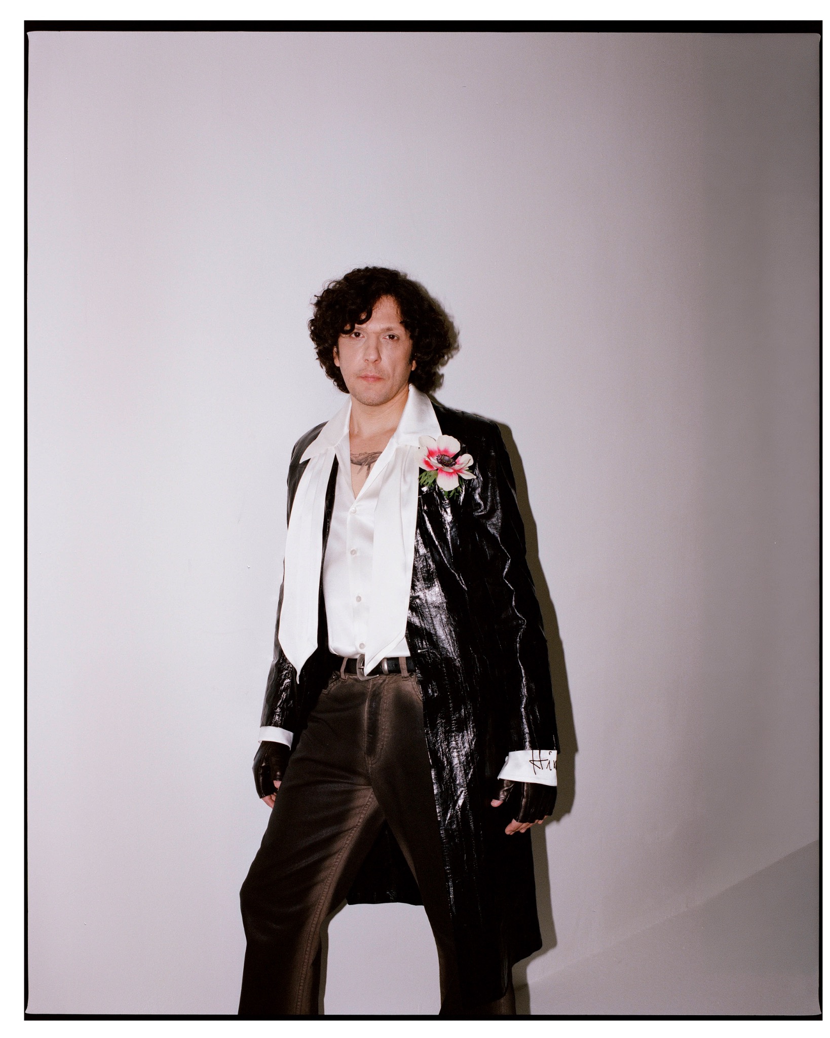 Ermal Meta standing in a black leather coat and white shirt, with a flower pinned to his lapel.