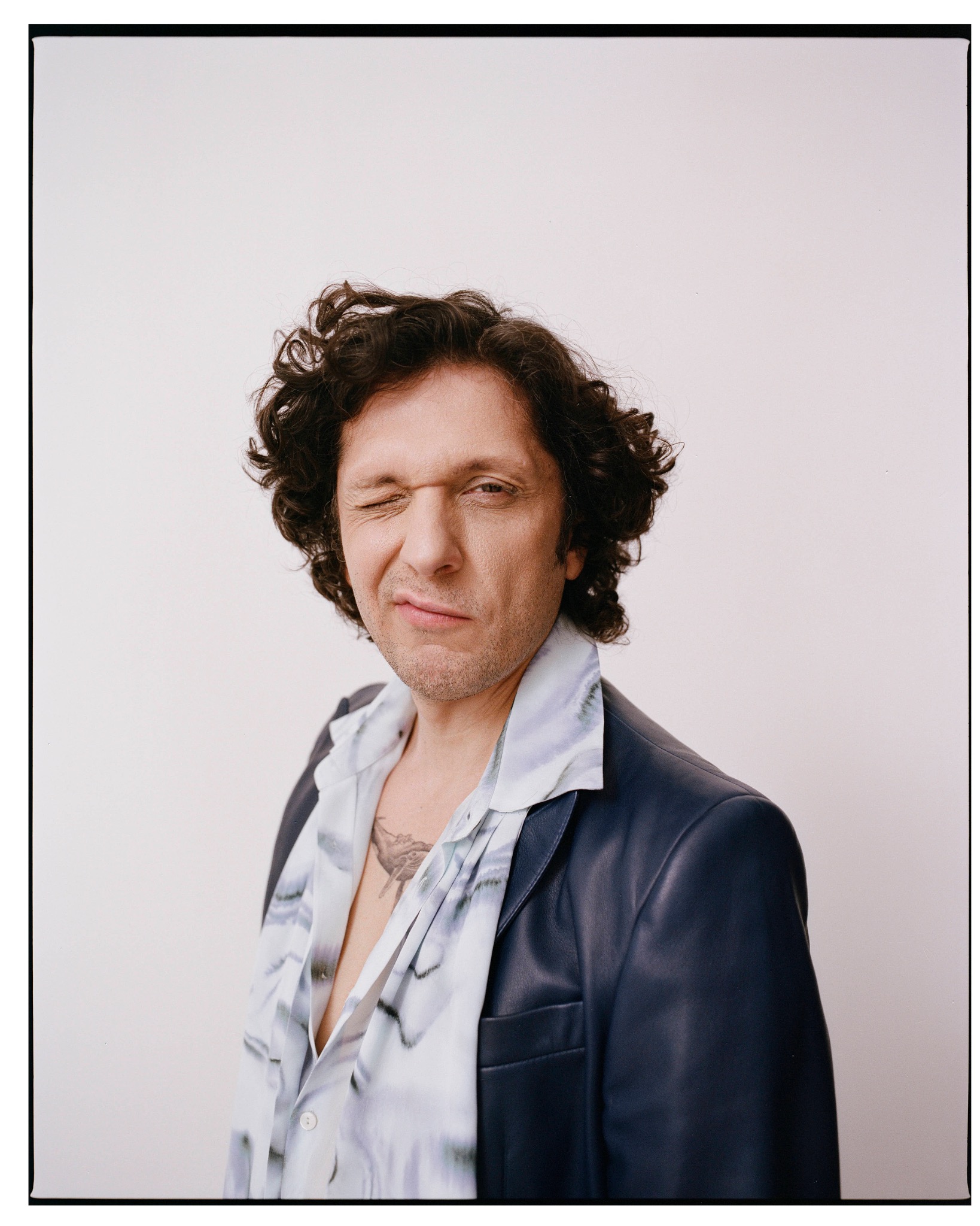 Portrait of Ermal Meta with his eyes closed, wearing a dark coat and a loosely tied patterned scarf.