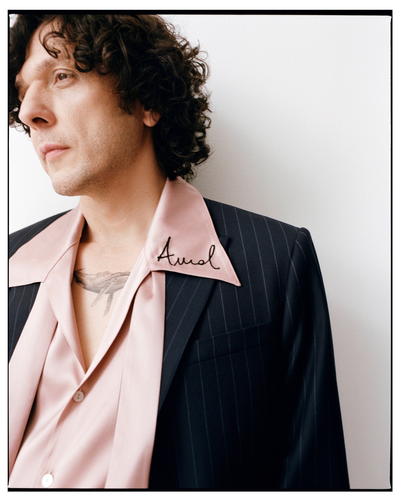 Close-up profile of Ermal Meta wearing a dark pinstriped jacket over a pink open-collar shirt.