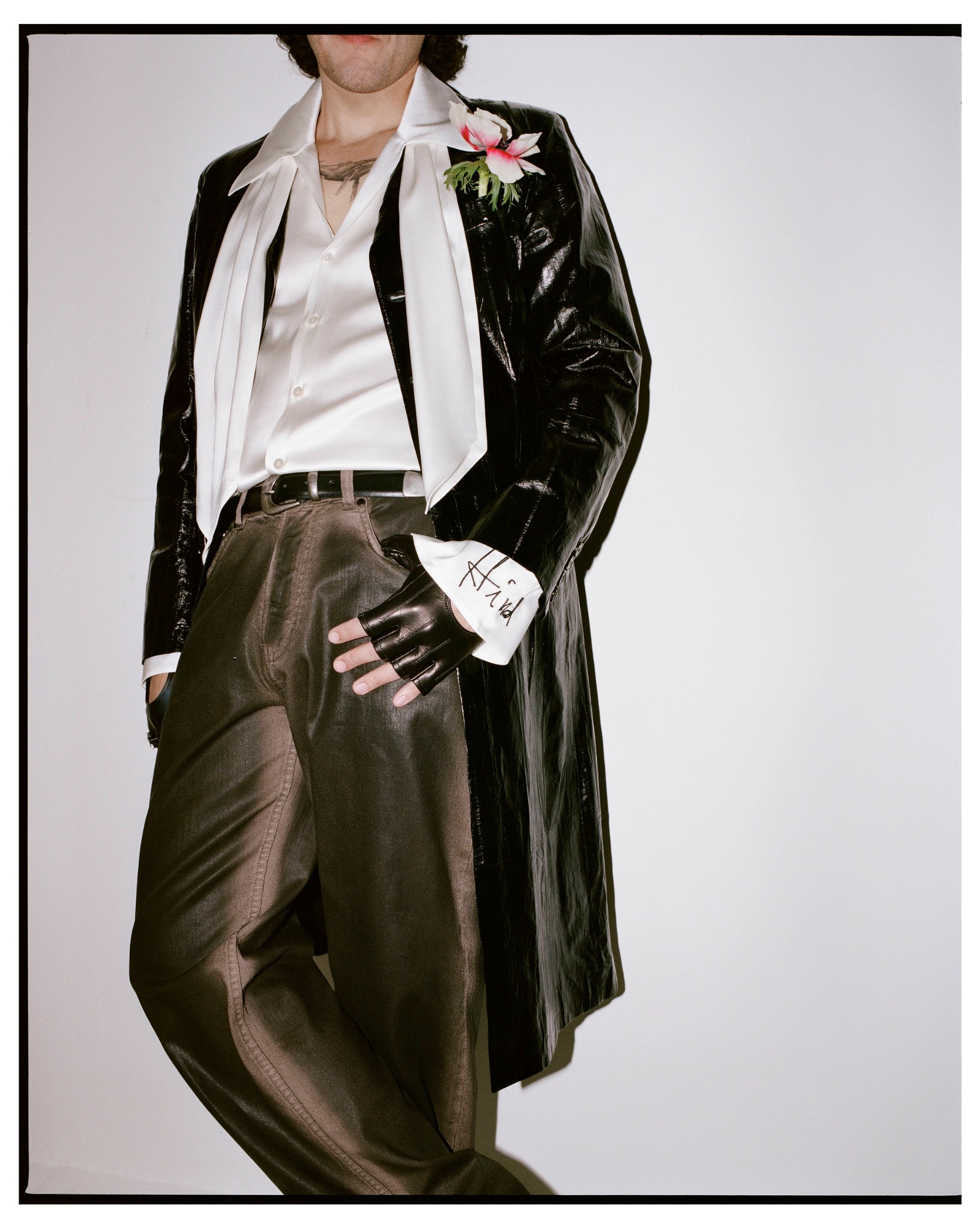 Cropped outfit detail of Ermal Meta in a black leather coat, white shirt, trousers and a flower on his lapel.