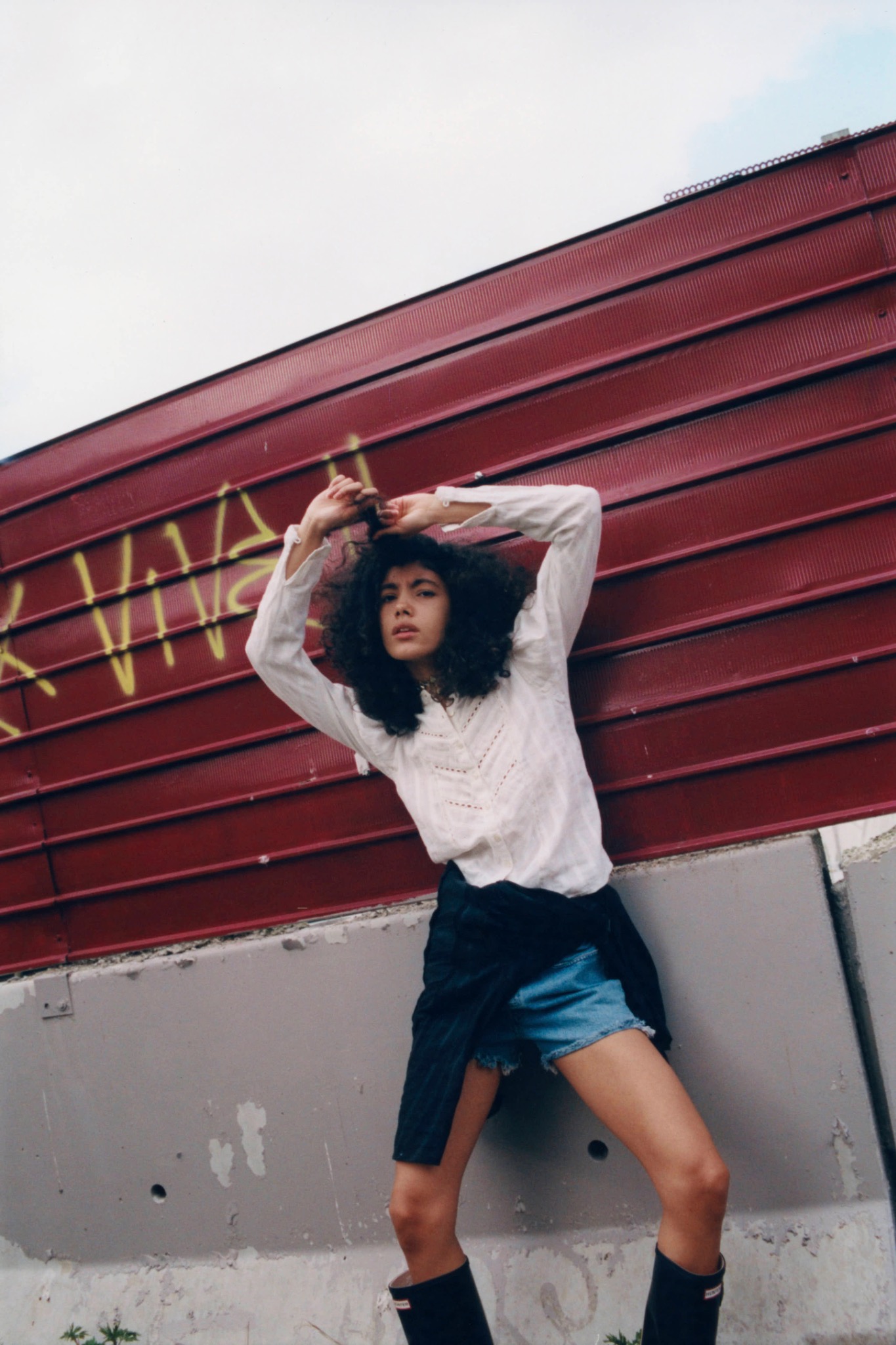 Curly-haired model raising both arms above their head beside a red wall, wearing a light top and layered shorts.