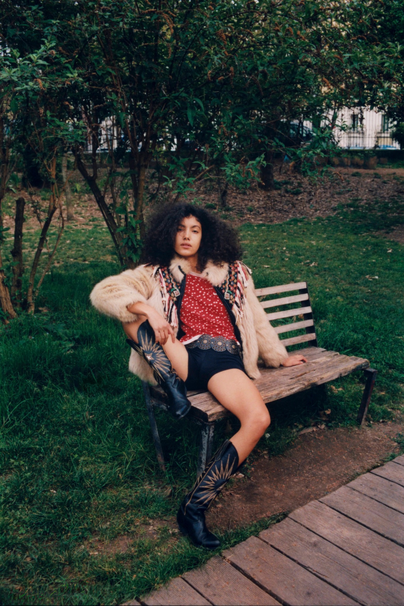Curly-haired model in a pale jacket and red top seated on a park bench, with trees behind.