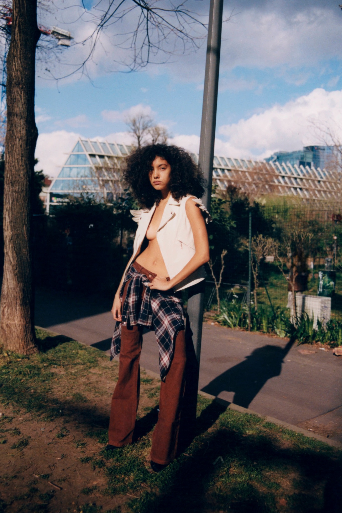 Curly-haired model in layered patterned clothing standing beside a tree and fence in a sunny park.