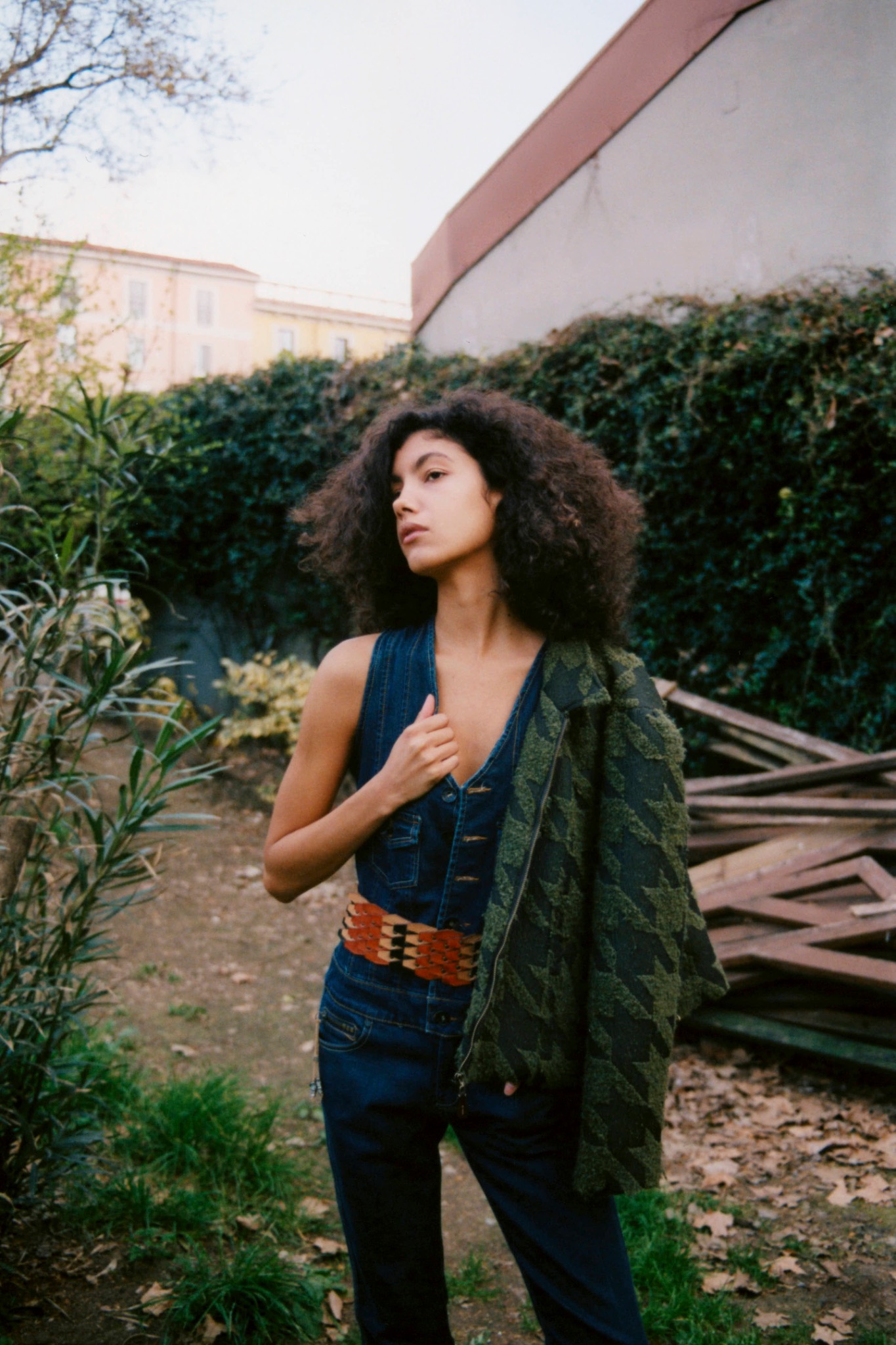 Curly-haired model holding a green patterned jacket over one shoulder outdoors, wearing a dark sleeveless top.