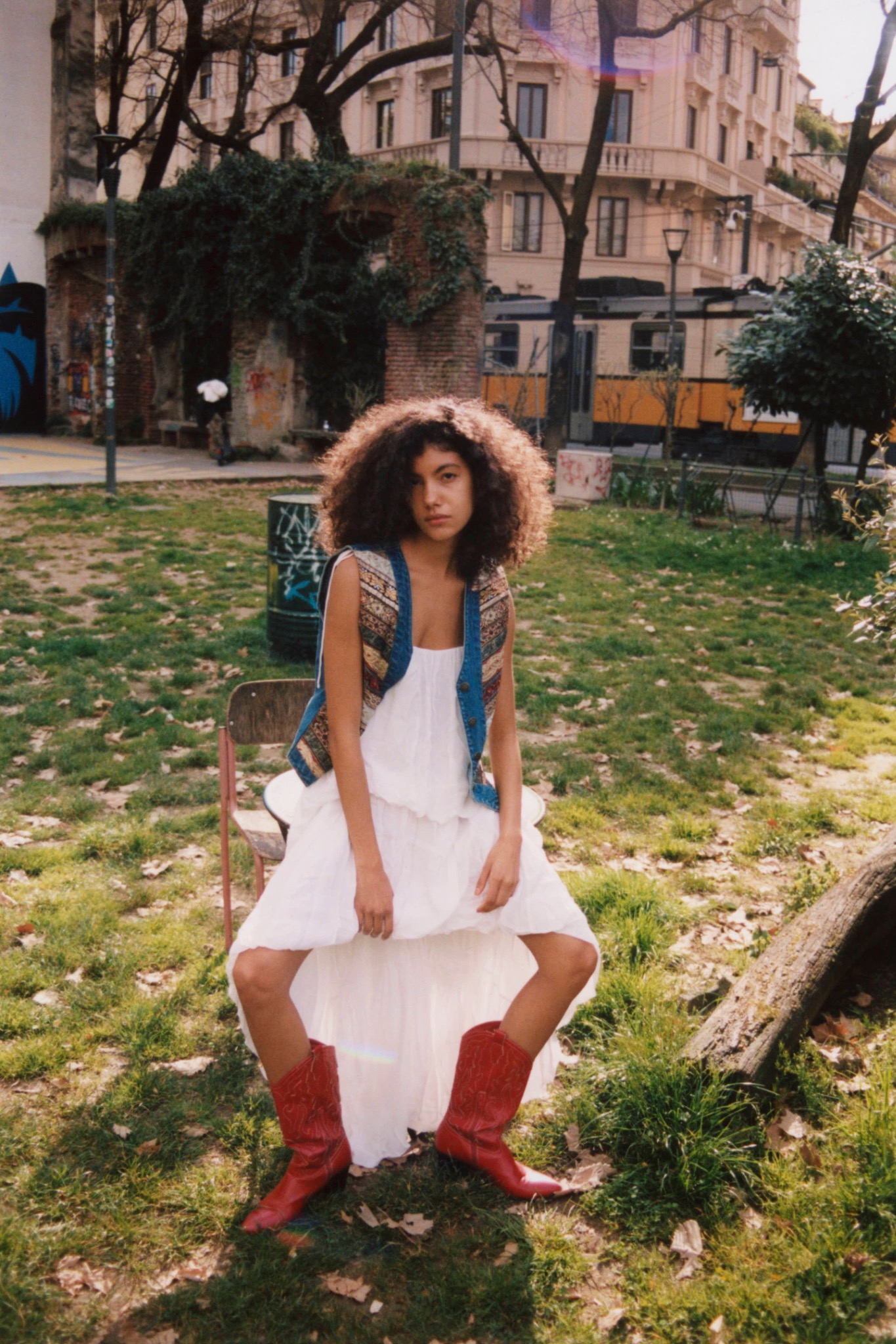 Curly-haired model seated on a low structure in a park, wearing a white skirt and red boots.