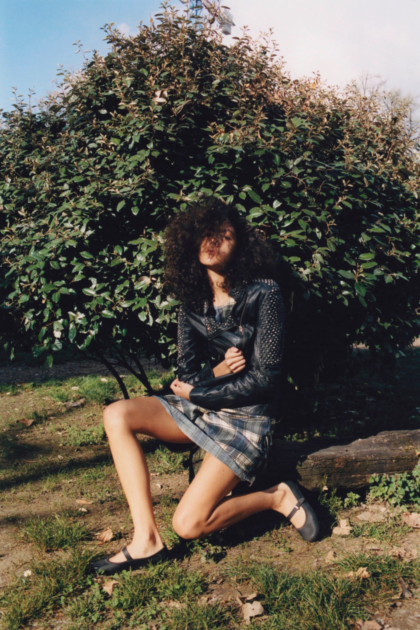 Curly-haired model seated beside flowering bushes in a dark patterned outfit.