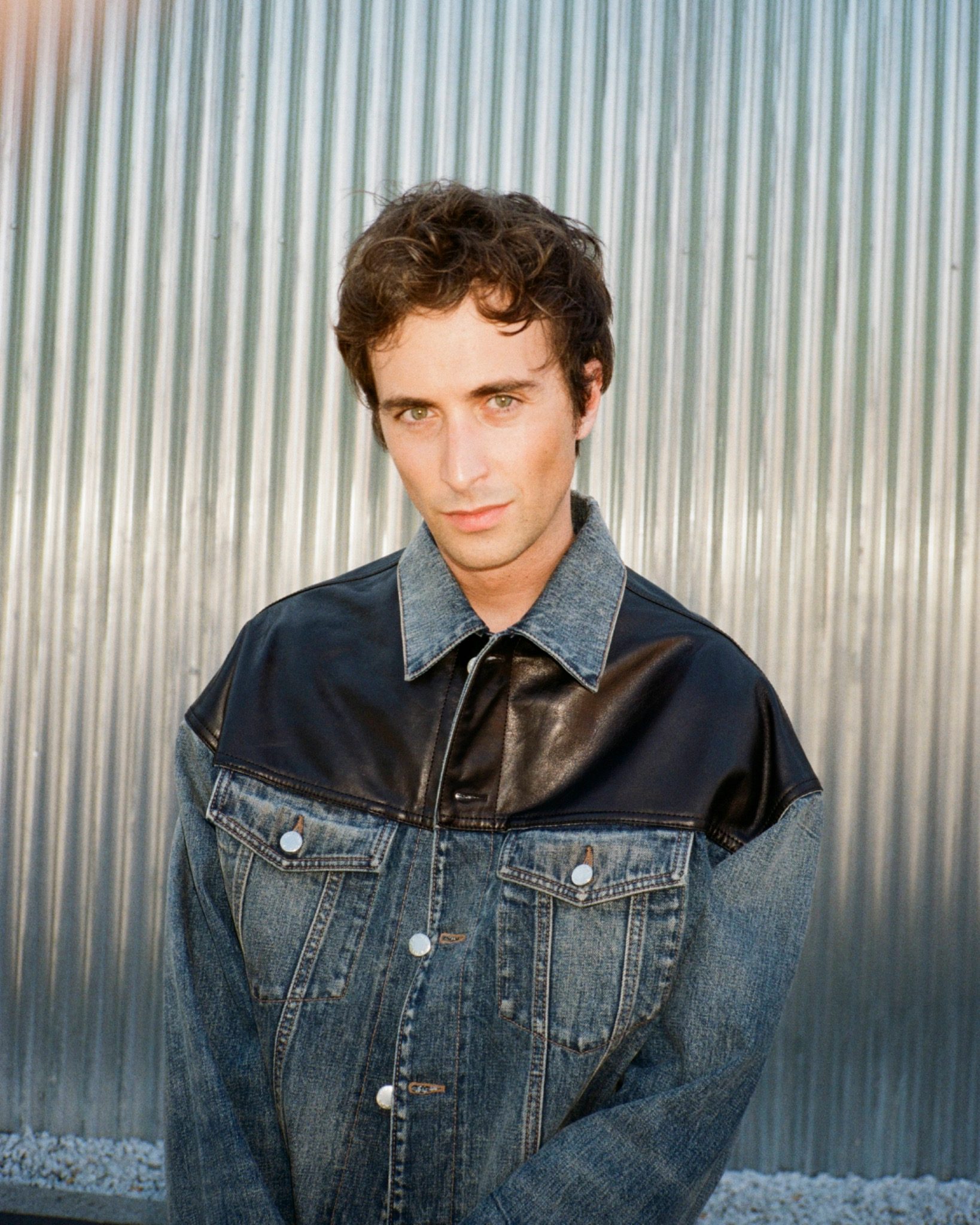 Portrait of Mattia in a denim jacket with a dark shoulder panel, standing against corrugated metal.