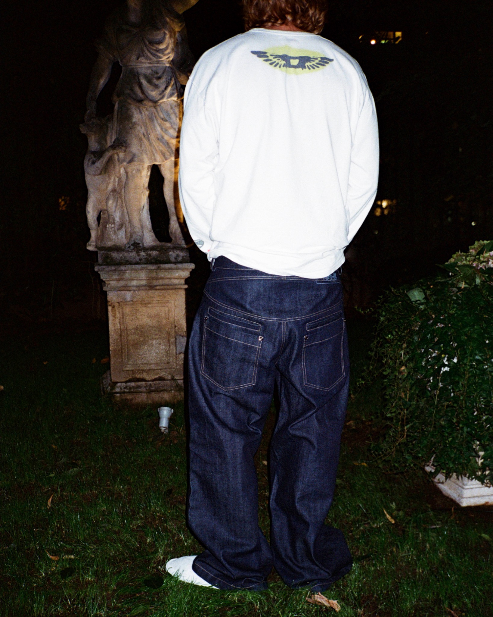 Jacopo seen from behind wearing a white long-sleeved top and dark jeans outdoors at night.