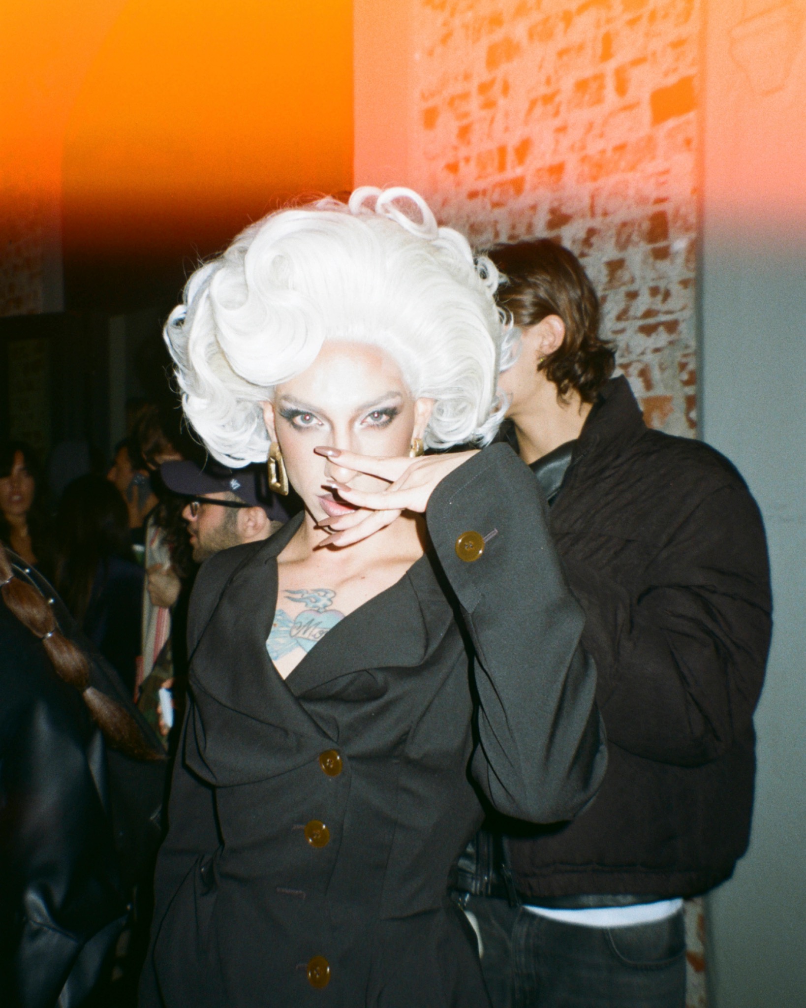 Angel in a white curled wig and a dark tailored outfit, holding a hand near their mouth in a warmly lit room.