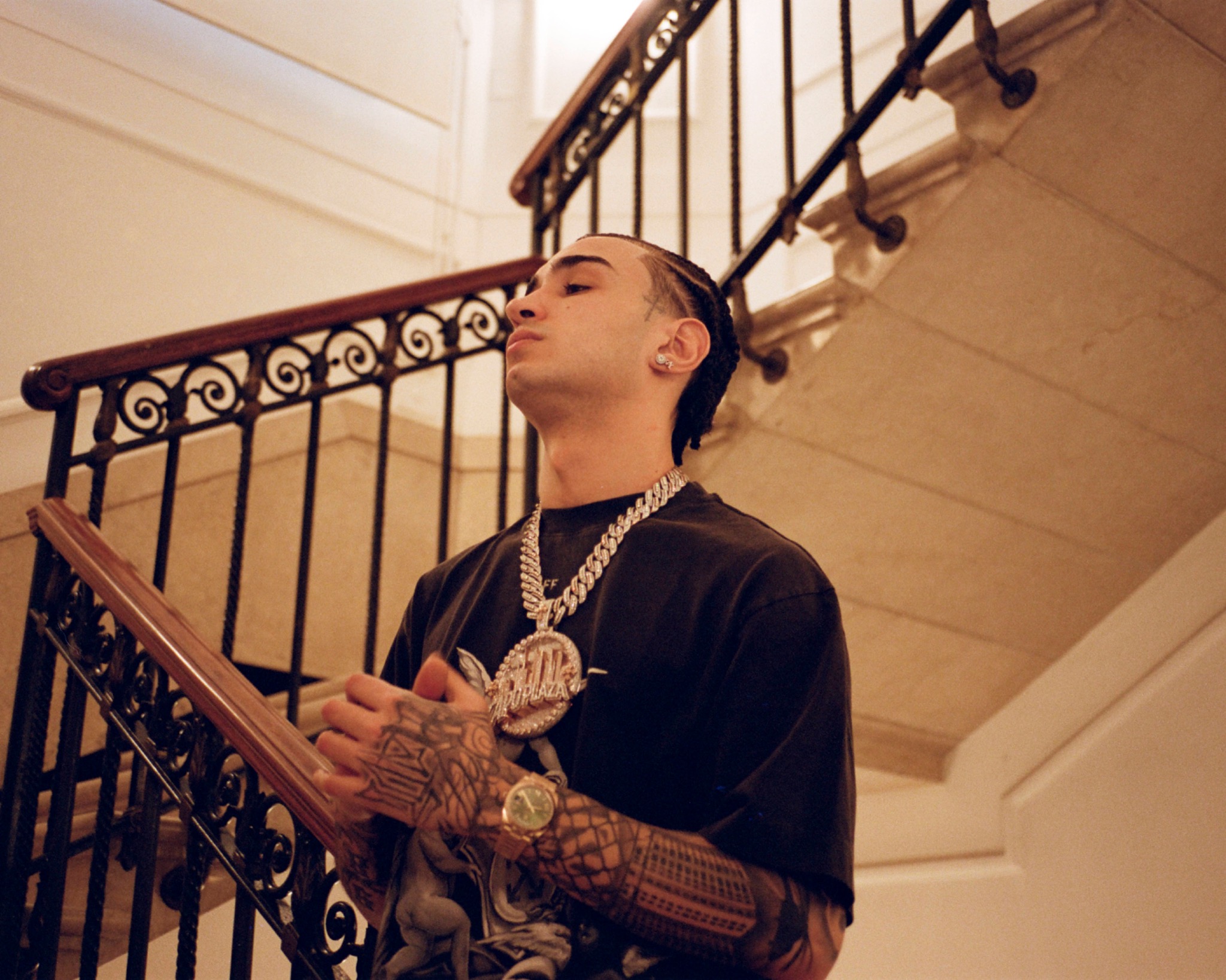Capo Plaza wearing jewellery and a dark printed T-shirt beside an ornate indoor staircase.