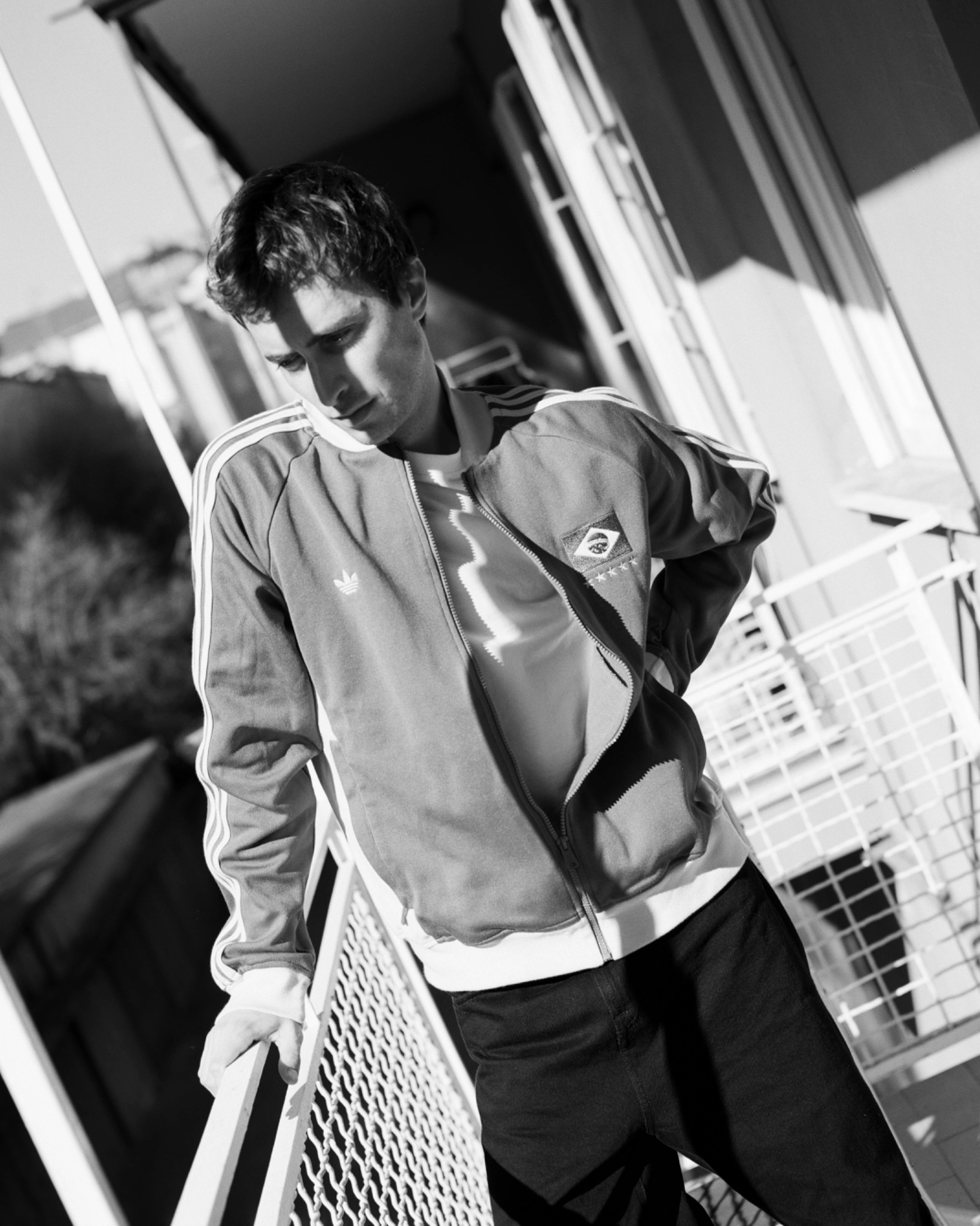 Black-and-white portrait of Mattia in a zip-up jacket beside an open doorway, lit by strong sunlight.