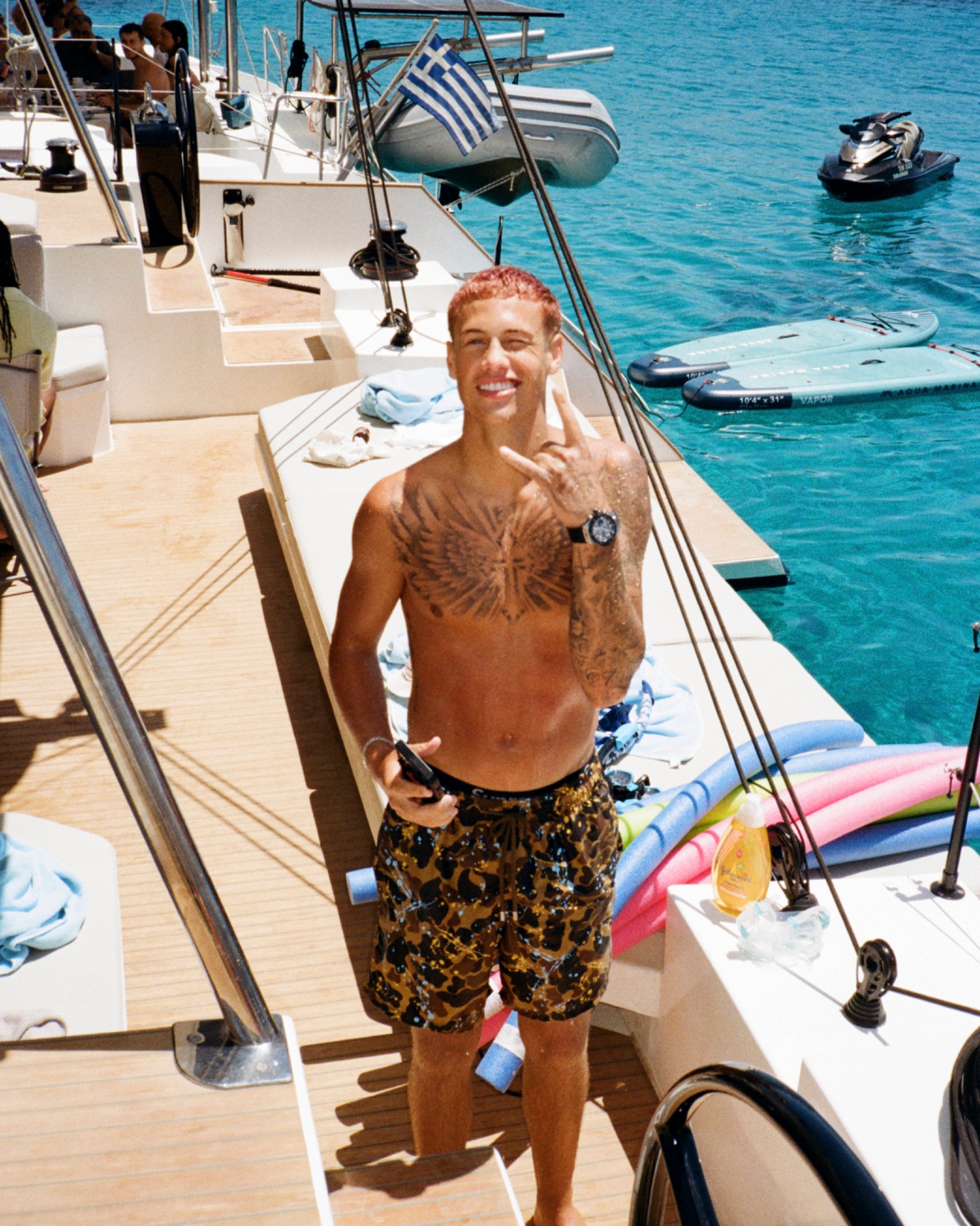 Matteo smiling on a sailboat in patterned shorts, with turquoise water and boats behind.