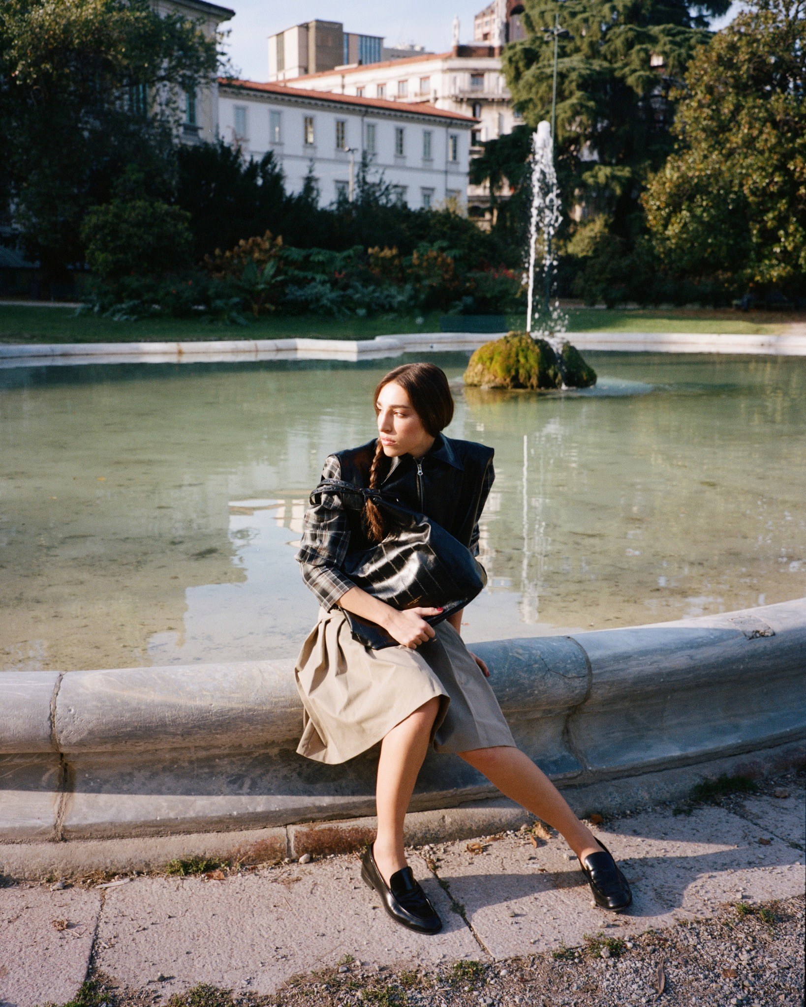 Ginevra seated beside a fountain in sunlight, wearing a checked jacket and skirt and holding a dark bag.