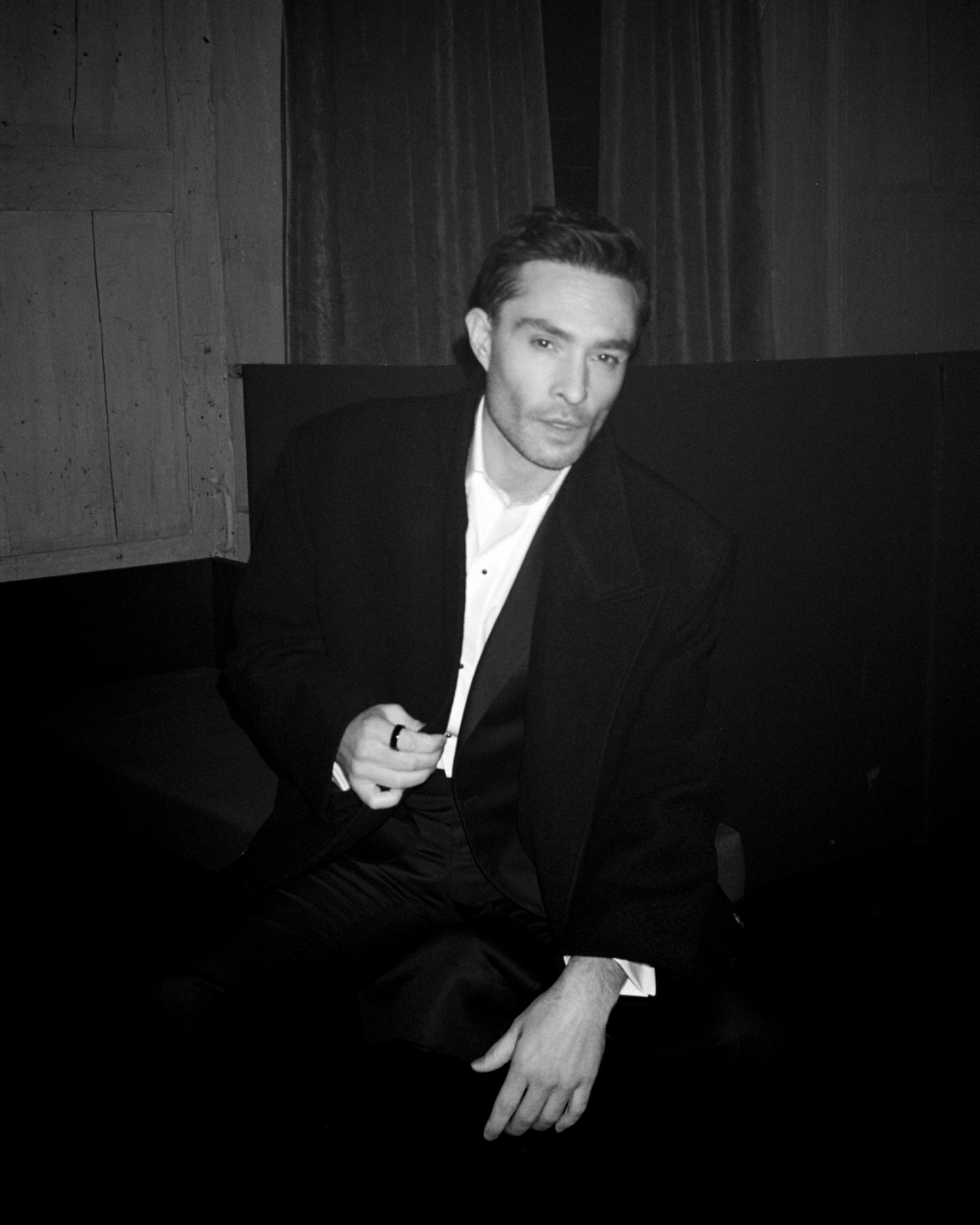 Black-and-white portrait of Ed seated in a dark suit and white shirt, holding a small object in one hand.