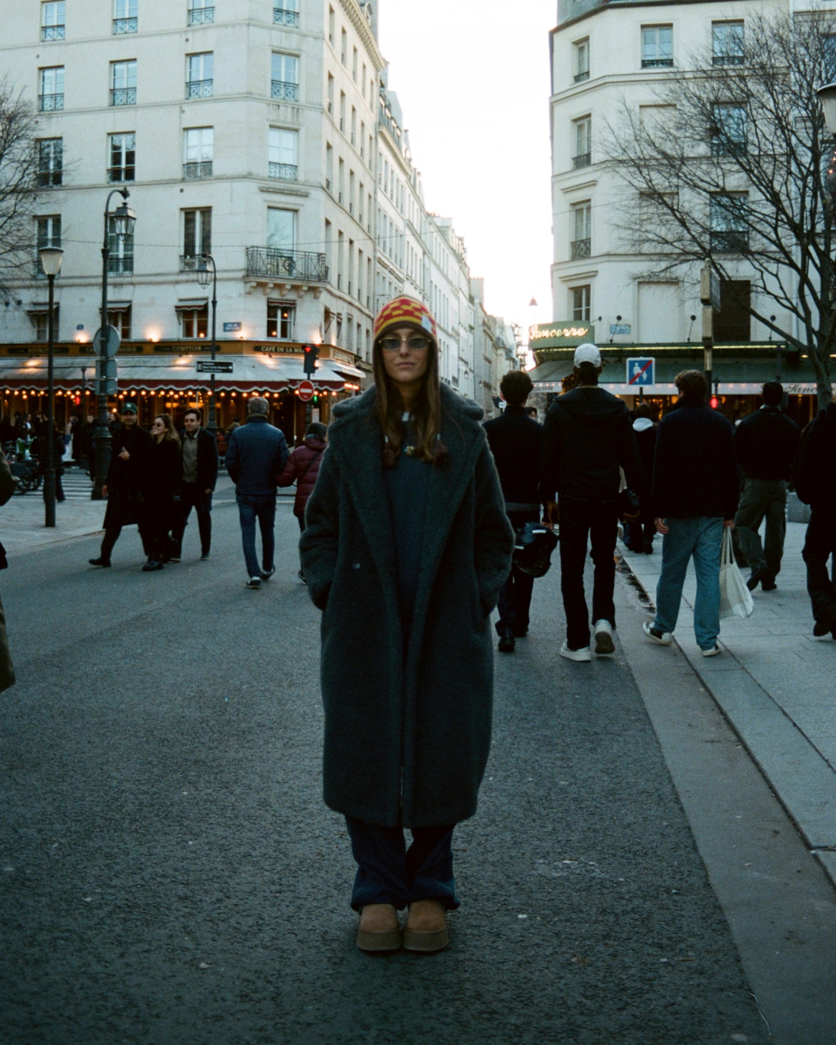 Full-length street portrait of Martina in a long dark coat, with pedestrians and tall city buildings behind.
