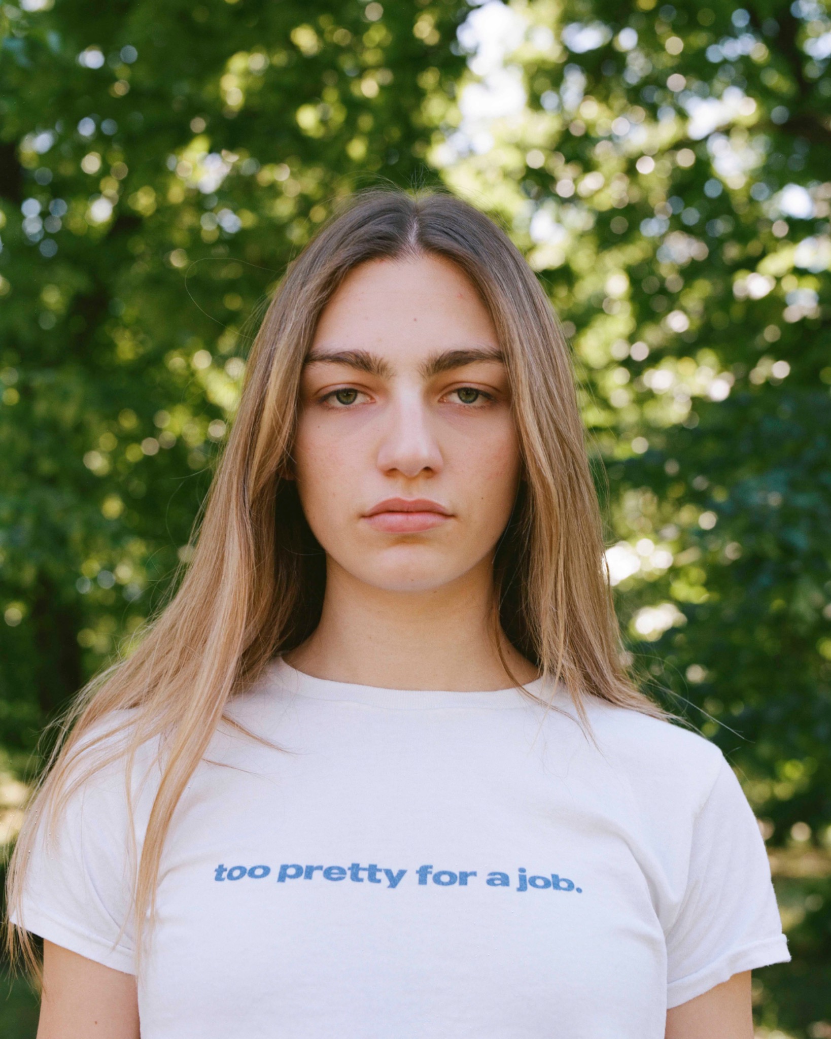 Outdoor portrait of Sara wearing a white T-shirt with blue lettering, leafy trees behind.