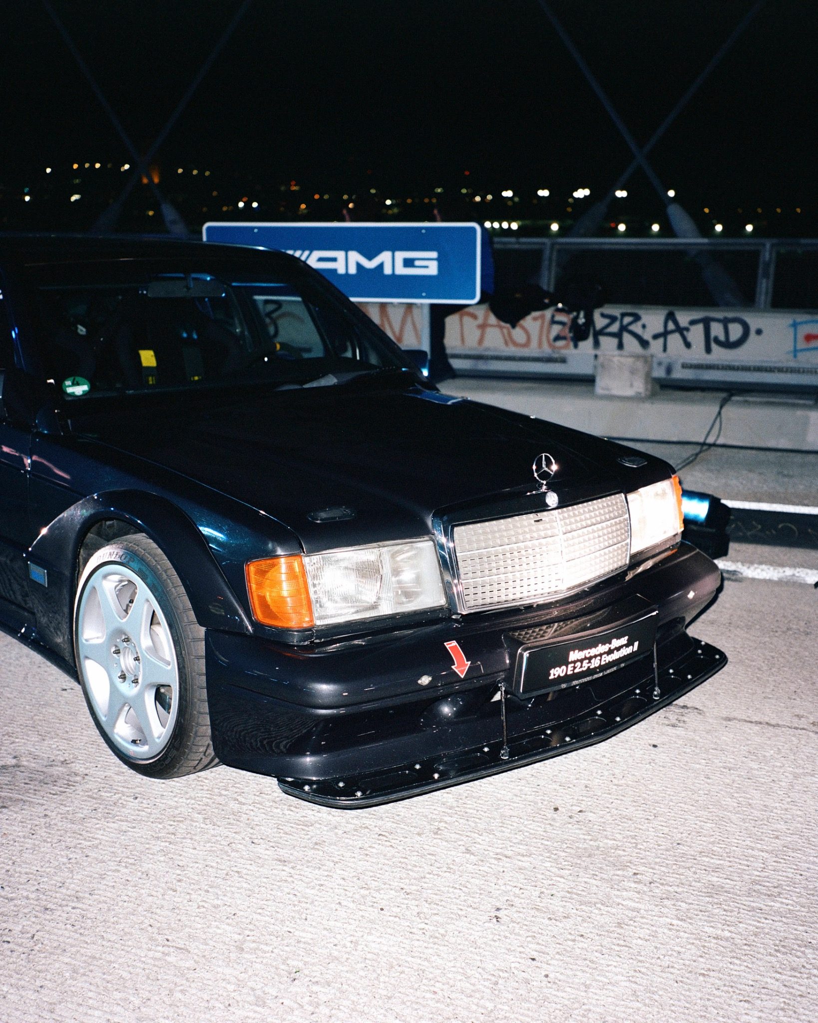 A black Mercedes parked at night, photographed from the front beneath lights and illuminated signage.