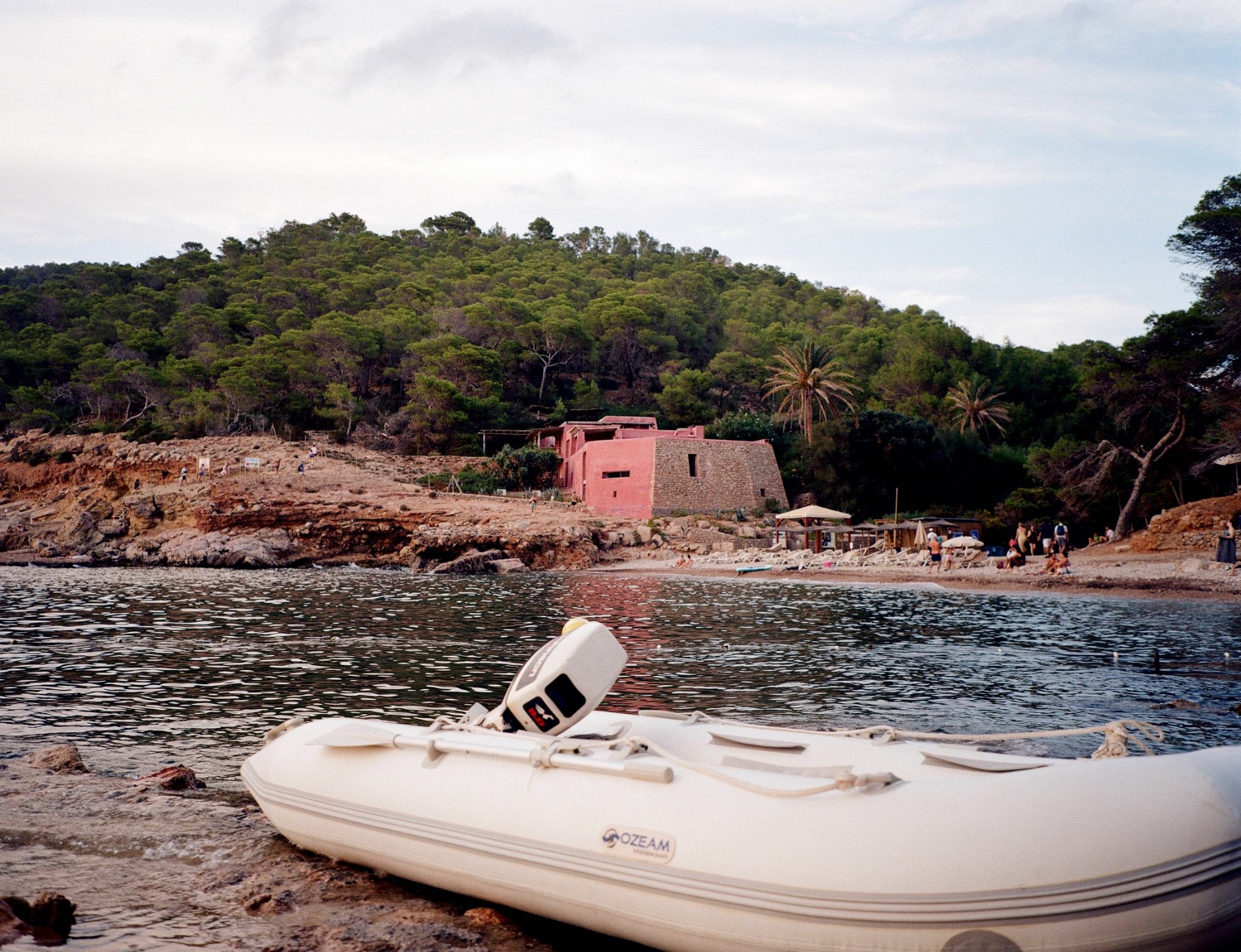 A white inflatable boat beside a rocky Ibiza shore, with a pink house among trees in the background.