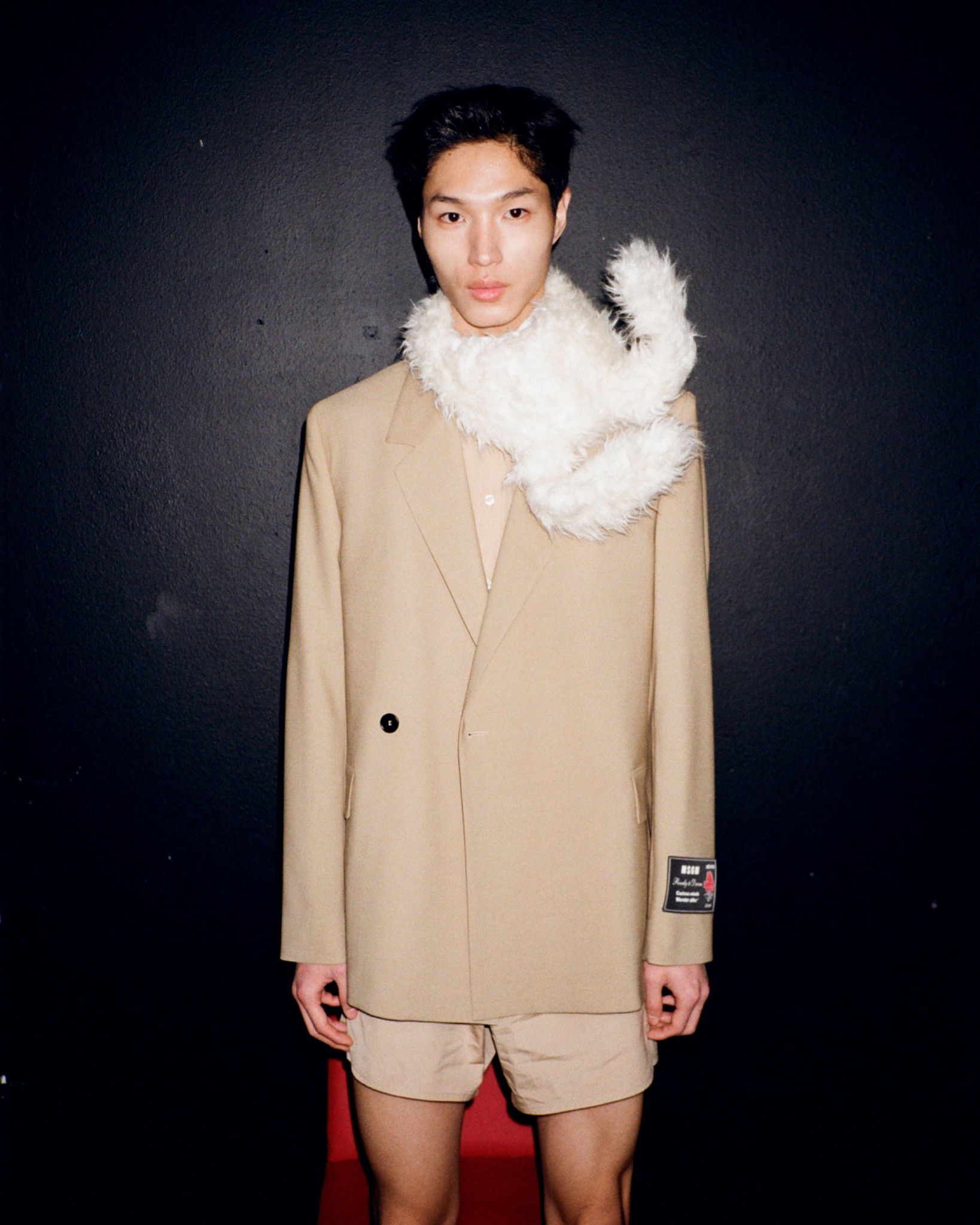Full-length portrait of a model in a beige jacket and shorts with a white fluffy scarf against a dark wall.