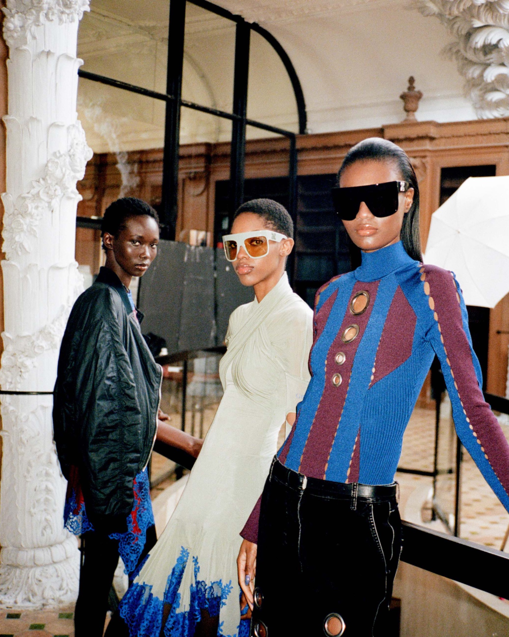 Farida, Jessie and Naki posing together indoors in colourful outfits and sunglasses, beside a decorative column.
