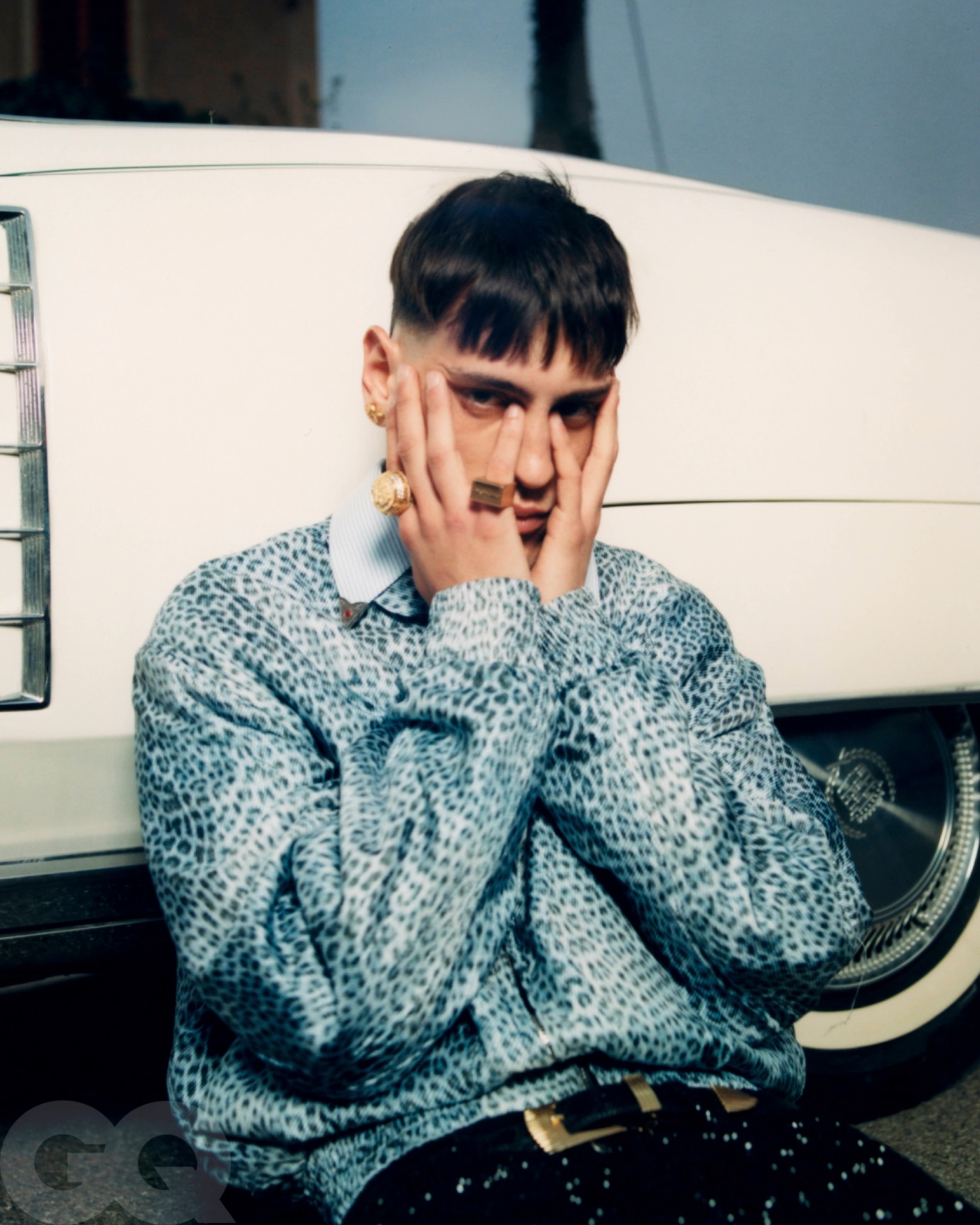 Promessa seated beside a white car with both hands against his cheeks, wearing a patterned knit sweater.