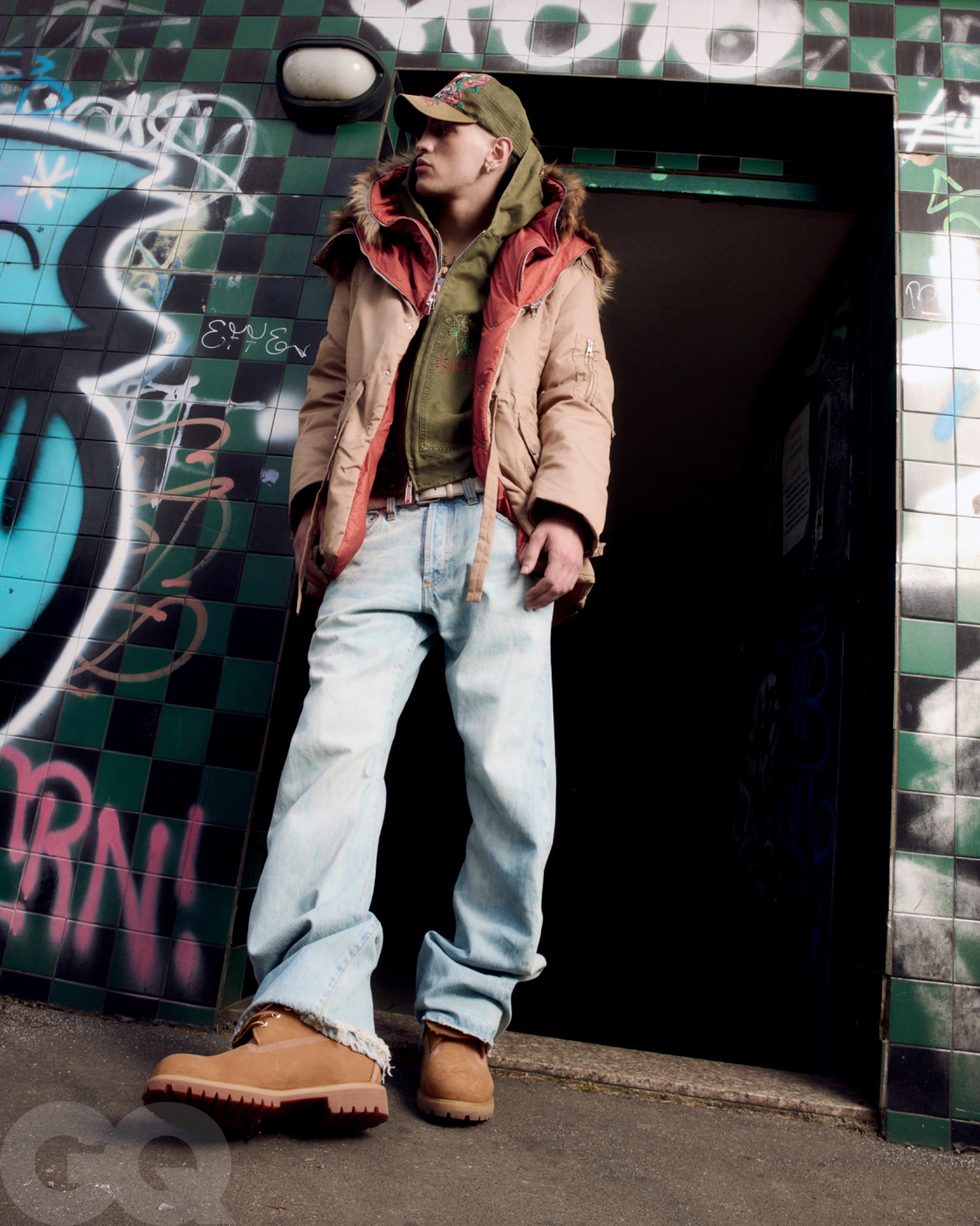 Low-angle portrait of Promessa in a cap, layered jackets and loose jeans standing in a graffiti-covered doorway.