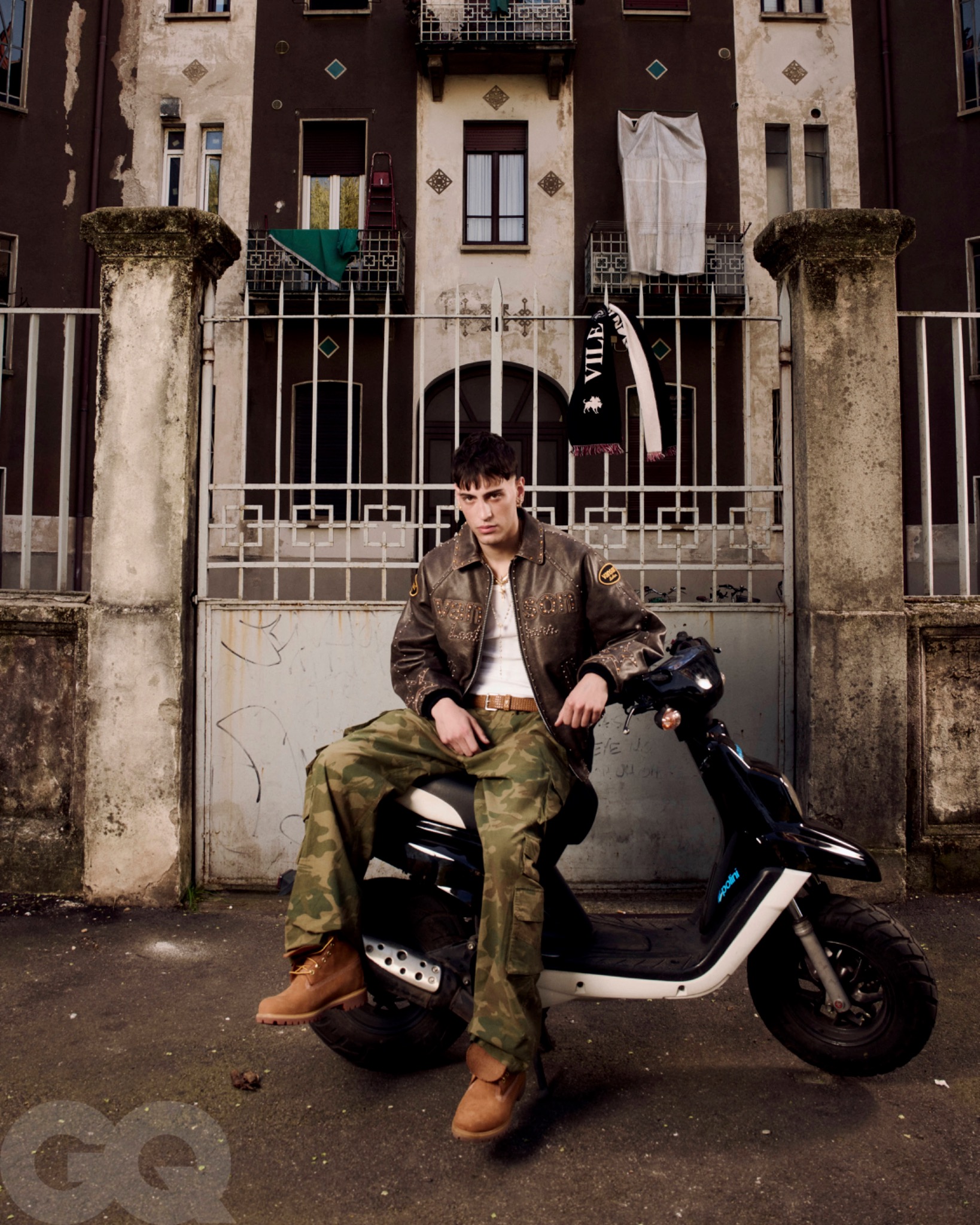 Promessa seated on a dark scooter outside an apartment building, wearing a jacket and camouflage trousers.