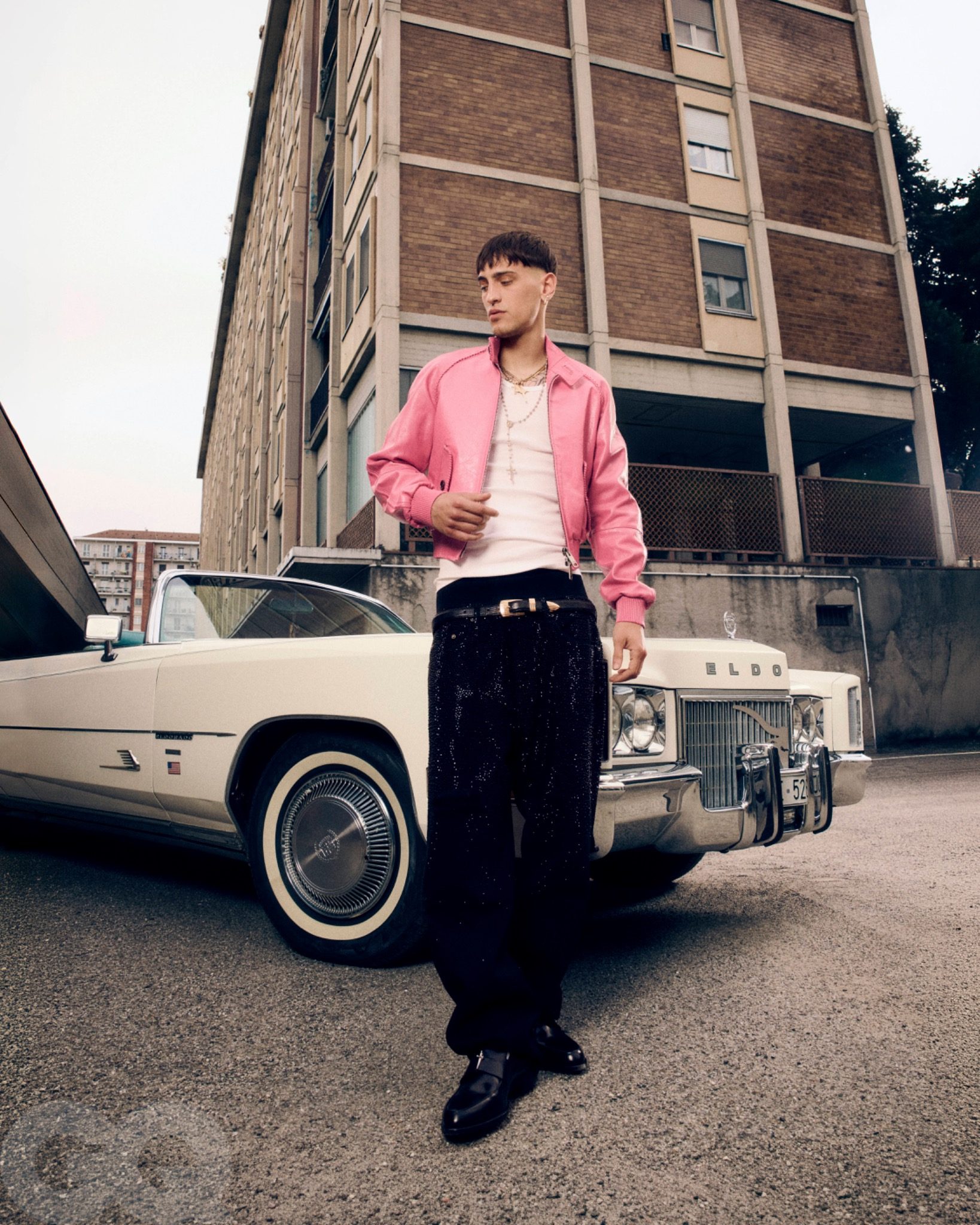 Promessa in a pink jacket standing beside a cream-coloured vintage car and a brick building.